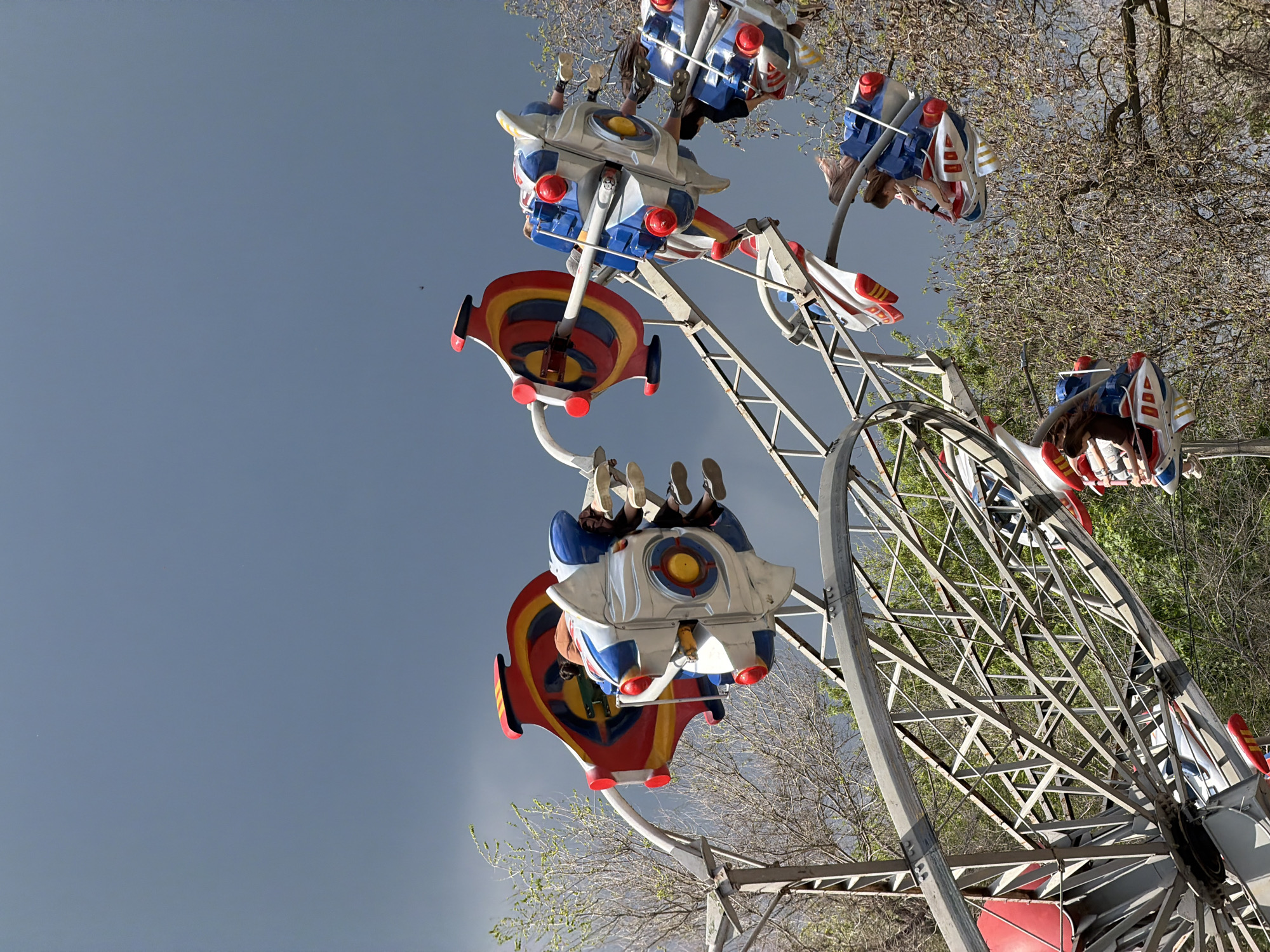The image shows an amusement park ride in Kamyshin, Russia. The ride features several small, airplane-shaped cars suspended from a central structure. People are seated in the cars, enjoying the ride. The structure is a metal frame with a circular track, and trees surround it. The sky is visible in the background.