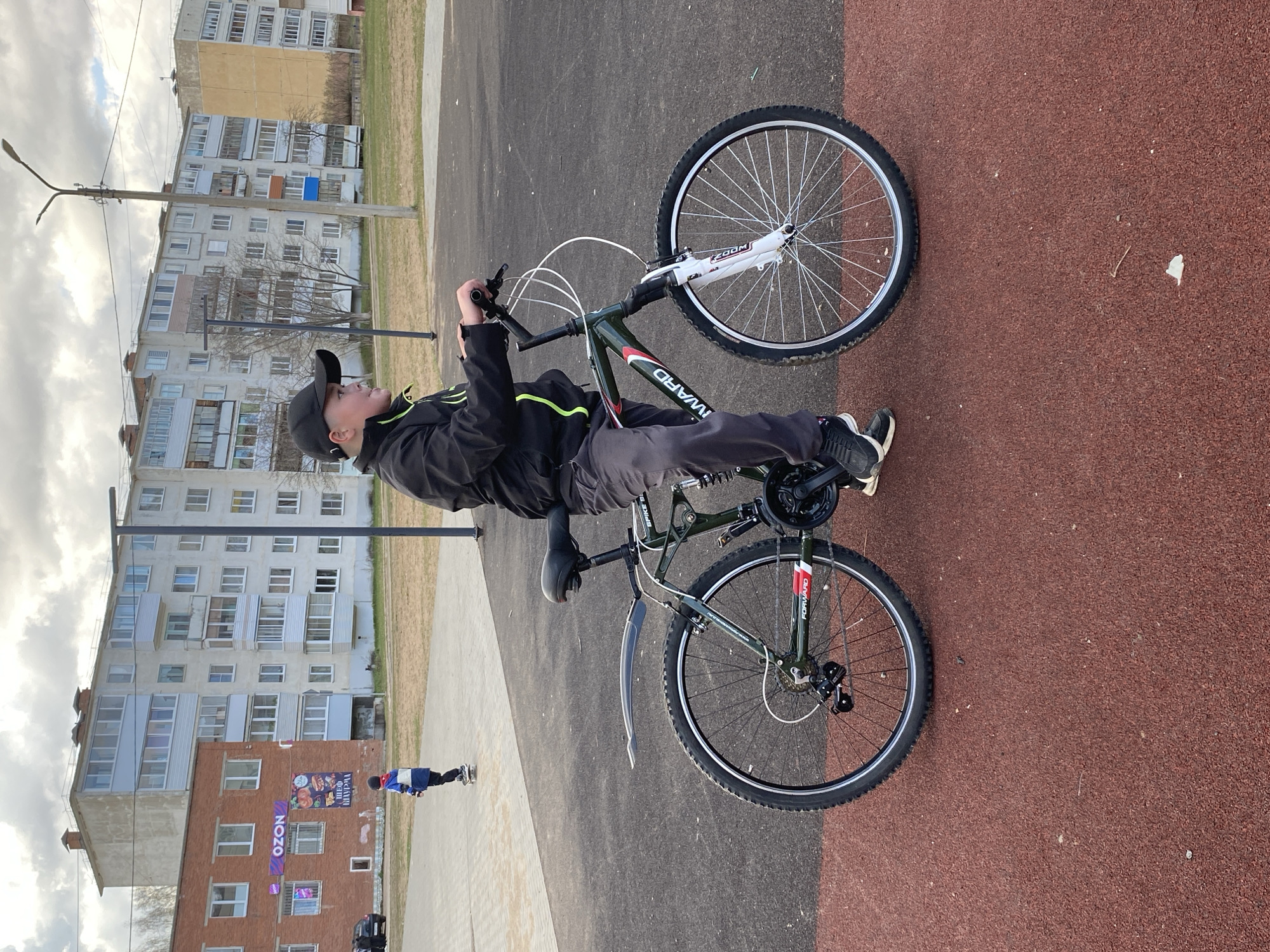 A young person, appearing male, is depicted outdoors in a semi-urban setting, positioned alongside a bicycle. The individual wears a black jacket with yellow accents, dark pants, and a black baseball cap. They are holding the handlebars of a dark green mountain bike, which features a white front suspension fork with "ZOOM" text, a "FORWARD" label on its frame (partially visible as "WARD"), and a front mudguard.
The scene takes place on a paved area, with a distinct reddish, granular surface (possibly a running track or sports court) to the right and a lighter grey pavement to the left. In the background, several multi-story residential apartment buildings of varying styles are visible, typical of a post-Soviet landscape. Some bare trees stand between the buildings and the paved area. A street lamp post is also visible. Another, smaller person in a blue and white jacket is seen in the distance near the buildings.
The activity appears to be a pause during or before a bike ride, with the subject standing next to the bicycle. The sky is overcast with a good amount of cloud cover, suggesting either a cloudy day or late afternoon.
Notable visible text includes "ZOOM" on the bicycle's front fork and "FORWARD" (partially "WARD") on the frame, indicating the brand. On one of the buildings in the background, a large "OZON" sign is prominently displayed, along with another partially visible sign with Cyrillic text, likely an advertisement for food (e.g., shashlik, sushi). The location context is Noviy, Russia, aligning with the visible signage and architecture.