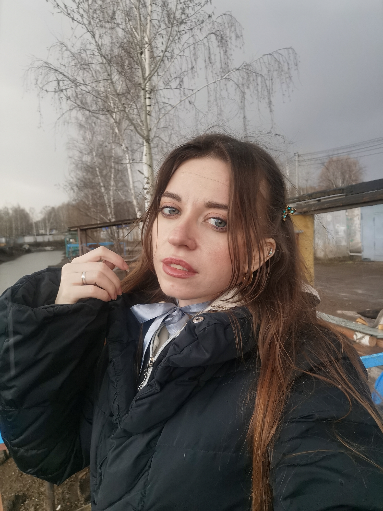 The image features a young woman outdoors, likely taking a selfie, against a backdrop of an overcast day. She has long, wavy brown hair, striking blue eyes, and a nose piercing on her right nostril. Her gaze is directed towards the camera with a focused expression, and her lips are slightly parted in a faint pout. She is dressed in a thick, dark-colored puffer jacket, with a lighter-colored garment visible underneath, accessorized by a light blue bow tied at the neck.
On her left hand, she wears a simple silver-toned ring on her ring finger and appears to be adjusting her jacket collar. Her left ear is adorned with multiple small stud earrings, while a decorative hair clip with green stones is placed in her hair above her right ear.
The background suggests a cool, possibly cold, season, such as late autumn or early spring, characterized by numerous deciduous trees with bare, intricate branches, including distinct white-barked birch trees. To her left, a dull, murky body of water, resembling a river or canal, stretches into the distance. To her right and further back, several utilitarian, possibly industrial or storage, buildings or garages are visible, indicating an urban or semi-urban environment. The sky is uniformly grey and overcast, and faint utility poles and wires can be seen in the distance. No visible text is present in the image.
