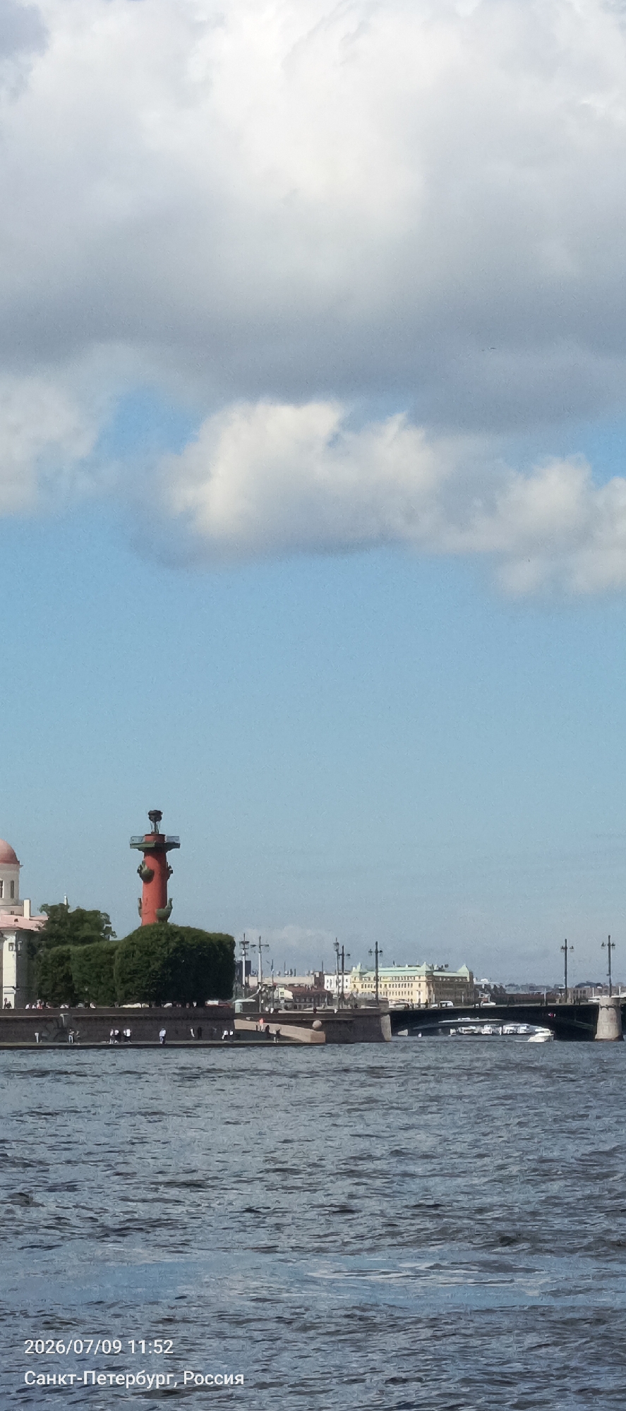 The image shows a waterfront scene in Saint Petersburg, Russia, likely from a boat. In the foreground is a body of water with ripples, suggesting a river or canal. On the far bank, a prominent red Rostral Column, a historical landmark and former lighthouse, stands on Vasilievsky Island. The column is adorned with figures and topped with a lantern. Adjacent to the column are manicured green bushes and a section of a quay with several people walking. Further along the embankment, there are buildings, streetlights, and a bridge spanning the water. The sky is partly cloudy with a mix of white and gray clouds against a bright blue background. A date and time stamp, "2026/07/09 11:52", and location text, "Санкт-Петербург, Россия", are visible in the bottom left corner.