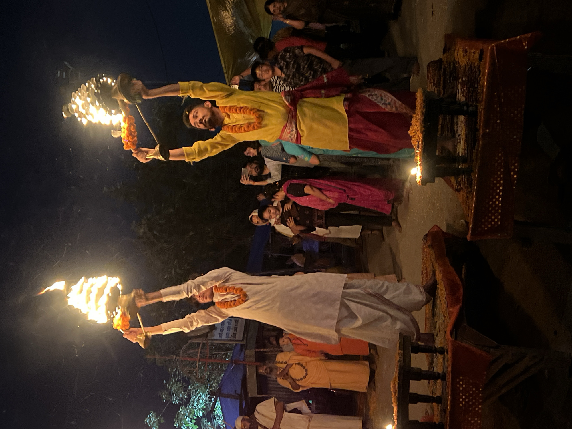 The image depicts a religious ceremony, likely an Aarti, taking place in Vrindavan, India, at night. Two individuals, dressed in traditional Indian attire, are holding aloft lamps with flames. The person at the top of the image is wearing a yellow kurta and red dhoti, adorned with a garland. The person at the bottom of the image is wearing a white kurta and dhoti, also with a garland. A crowd of onlookers is gathered around, some of whom are holding up their phones to capture the scene. A small stand with lit candles is to the right in the picture. The lighting is low, with the flames providing the primary source of illumination. The atmosphere appears reverent and festive.