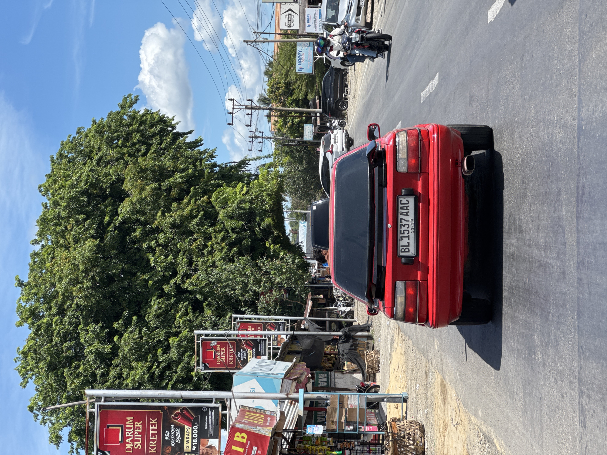 The image shows a street scene in Tumpok, Indonesia. In the foreground, a red car with license plate BL 1537 AAC is driving on a paved road. The car has a spoiler on the back and a visible exhaust pipe. On the left side of the road, there are several shops and stalls, including one with a sign for "Djarum Super Kretek". A large tree with lush green foliage dominates the background, with a bright blue sky and scattered white clouds above. Further down the road, other vehicles, including cars and motorcycles, are visible, as are power lines and utility poles. One motorcycle has two people riding on it. The overall impression is a sunny day in a bustling urban or semi-urban environment.