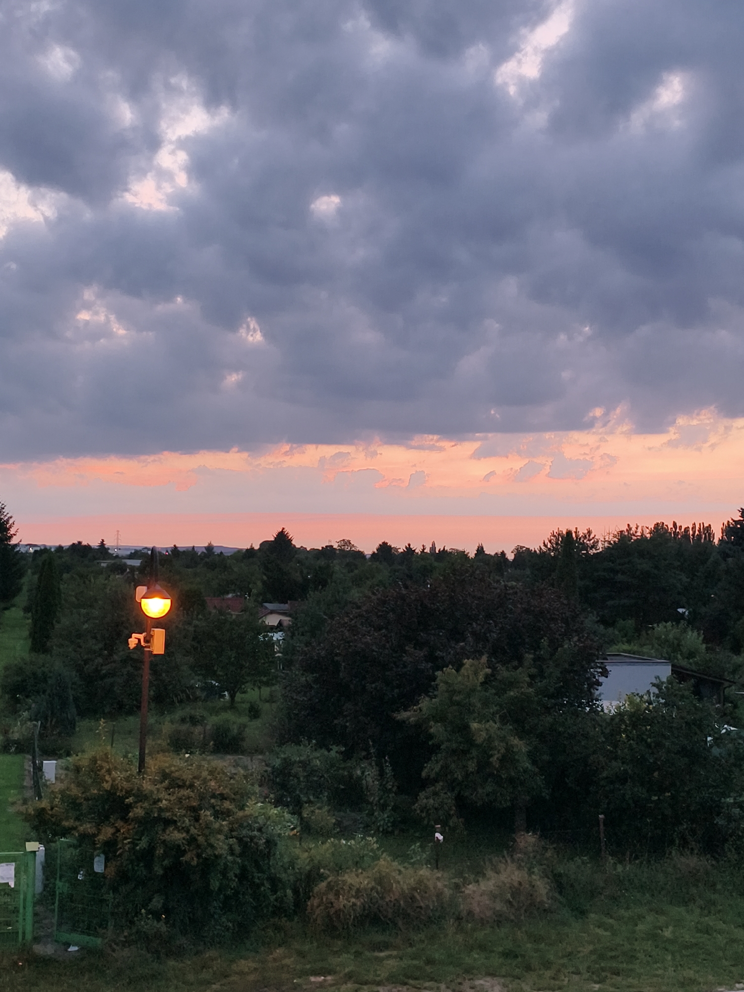The image captures a view of the outskirts of Tczew, Poland, during sunset. The sky is filled with dramatic, dark grey clouds, with a band of soft pink and orange hues visible along the horizon, indicative of the setting sun. In the foreground, a lamppost with an illuminated orange light stands prominently, casting a warm glow. The lamppost is mounted on a pole and has a small camera attached to it. Surrounding the lamppost and extending into the midground is dense foliage, consisting of various trees and bushes in shades of green and dark red. A glimpse of residential buildings and a white structure, possibly a shed or garage, can be seen peeking through the trees. In the far distance, a faint outline of power lines and transmission towers suggest the presence of more developed areas beyond the immediate landscape. The overall scene depicts a semi-rural environment transitioning into evening.