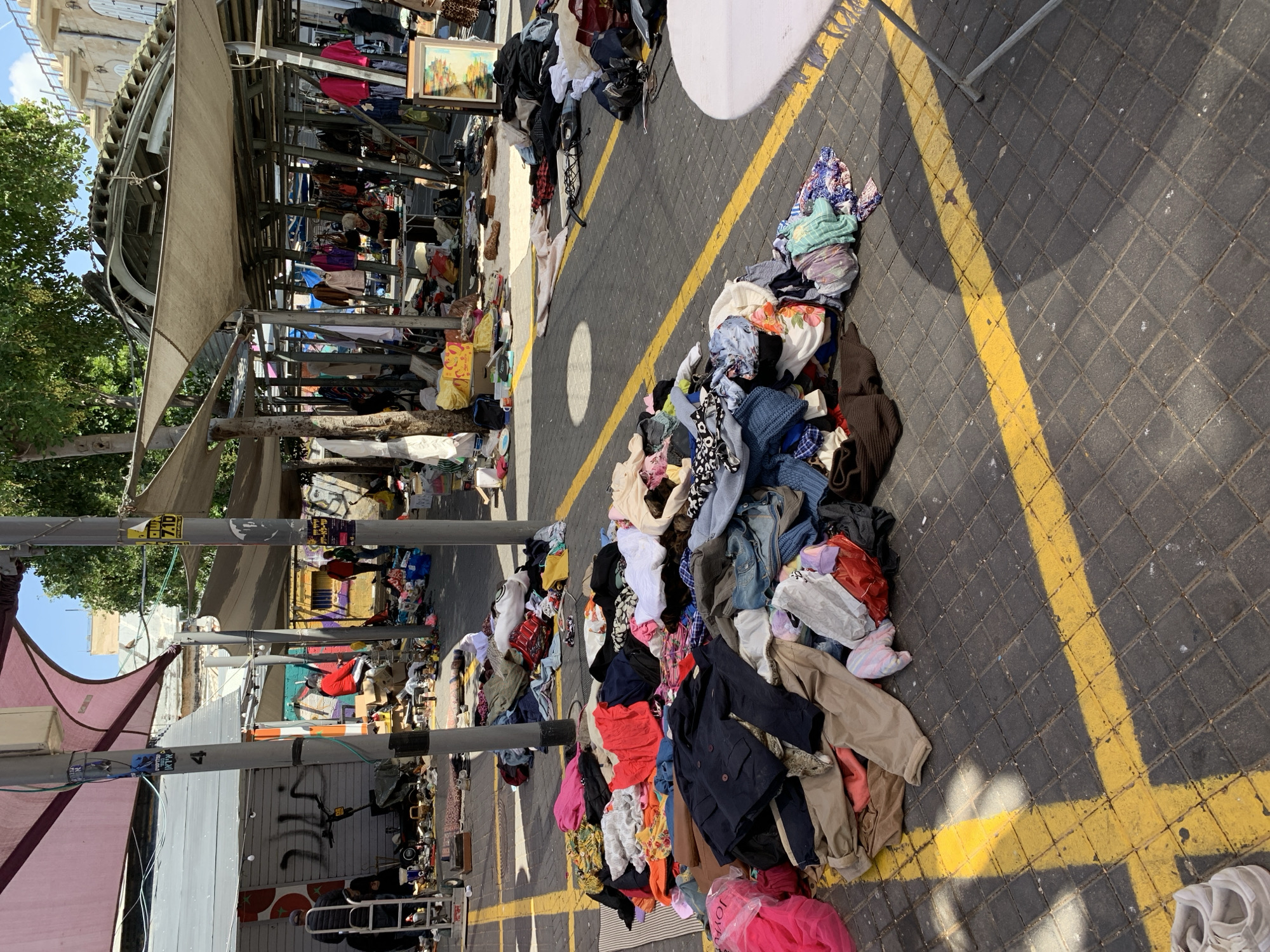 The image depicts a street scene in Tel Aviv, Israel, likely within the Carmel Market (Shuk HaCarmel) area. A narrow paved pedestrian zone is marked with thick yellow lines. Large piles of used clothing are heaped directly onto the pavement along these lines. Vendors sell additional clothing from stalls with metal framing and awnings to the left of the piles. Other stalls and buildings can be seen farther down the street.