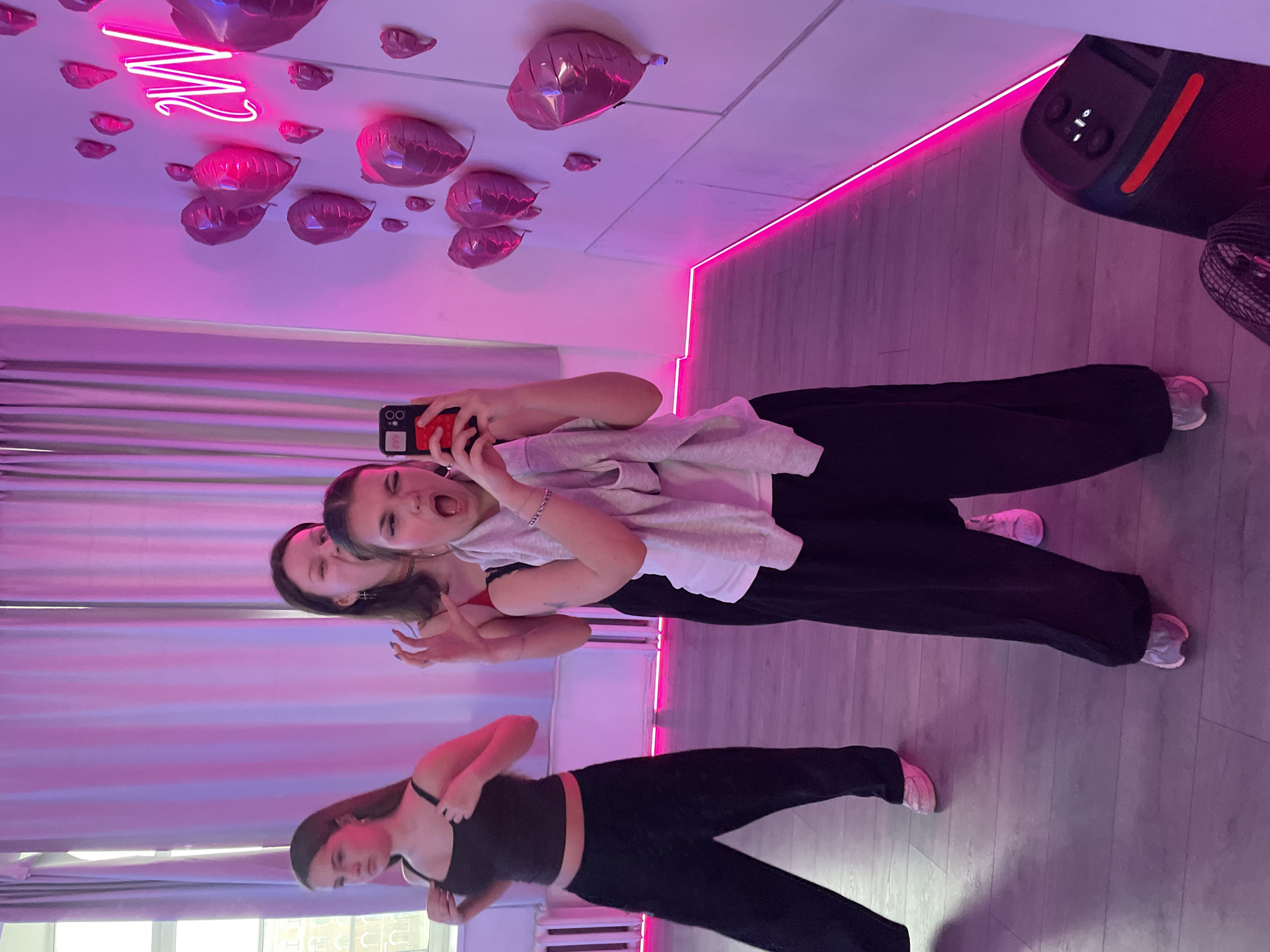 This image depicts two young women taking a mirror selfie in a vibrant, neon-lit room, likely located in Saint Petersburg, Russia. The room is characterized by strong artificial pink and purple lighting, emanating from LED strips along the walls and a prominent "M" (or "W") shaped neon sign affixed to the white ceiling.
In the foreground, one young woman holds a smartphone with a bright red case, directing it towards a large mirror to capture the reflection of herself and her companion. She makes a playful and energetic expression, with her mouth open and eyes wide. She is dressed in a light grey long-sleeved top over a white garment and dark, loose-fitting pants. Behind her, another woman is partially visible, looking towards the camera with a subtle smile.
The mirror's reflection also shows a third young woman, dressed in a black sports bra or tank top and black pants, adjusting her attire. The ceiling is decorated with numerous shiny pink heart-shaped foil balloons. The floor is made of light-toned wooden planks. In the upper right portion of the image, a black portable speaker with an orange accent stripe and illuminated control buttons is visible, along with part of a black fan. All individuals are dressed in casual athletic wear, including sneakers. The scene evokes a lively and dynamic atmosphere, suggesting a recreational or practice space, possibly a dance studio, during the evening or night due to the prominent artificial lighting. The phone case has some partially obscured text.