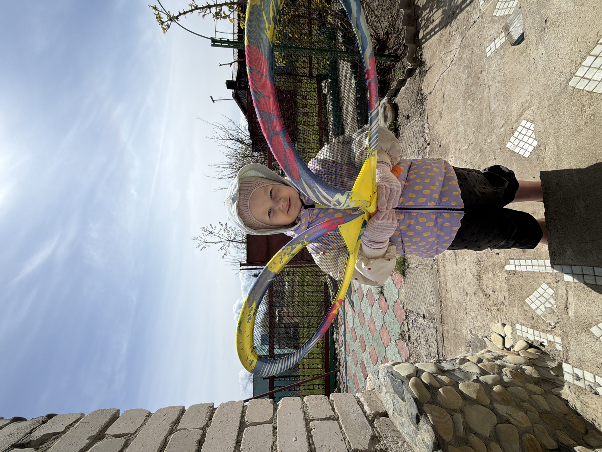 A young child stands outdoors, holding a large badminton racket. The child wears a light-colored hooded hat, a light purple jacket with yellow polka dots, dark pants, and gloves. The badminton racket is colorful with yellow, blue, and red accents. The background includes a brick wall, sky, a building, and a textured ground surface with both concrete and patterned tiles. The location is Kamyshin, Russia.