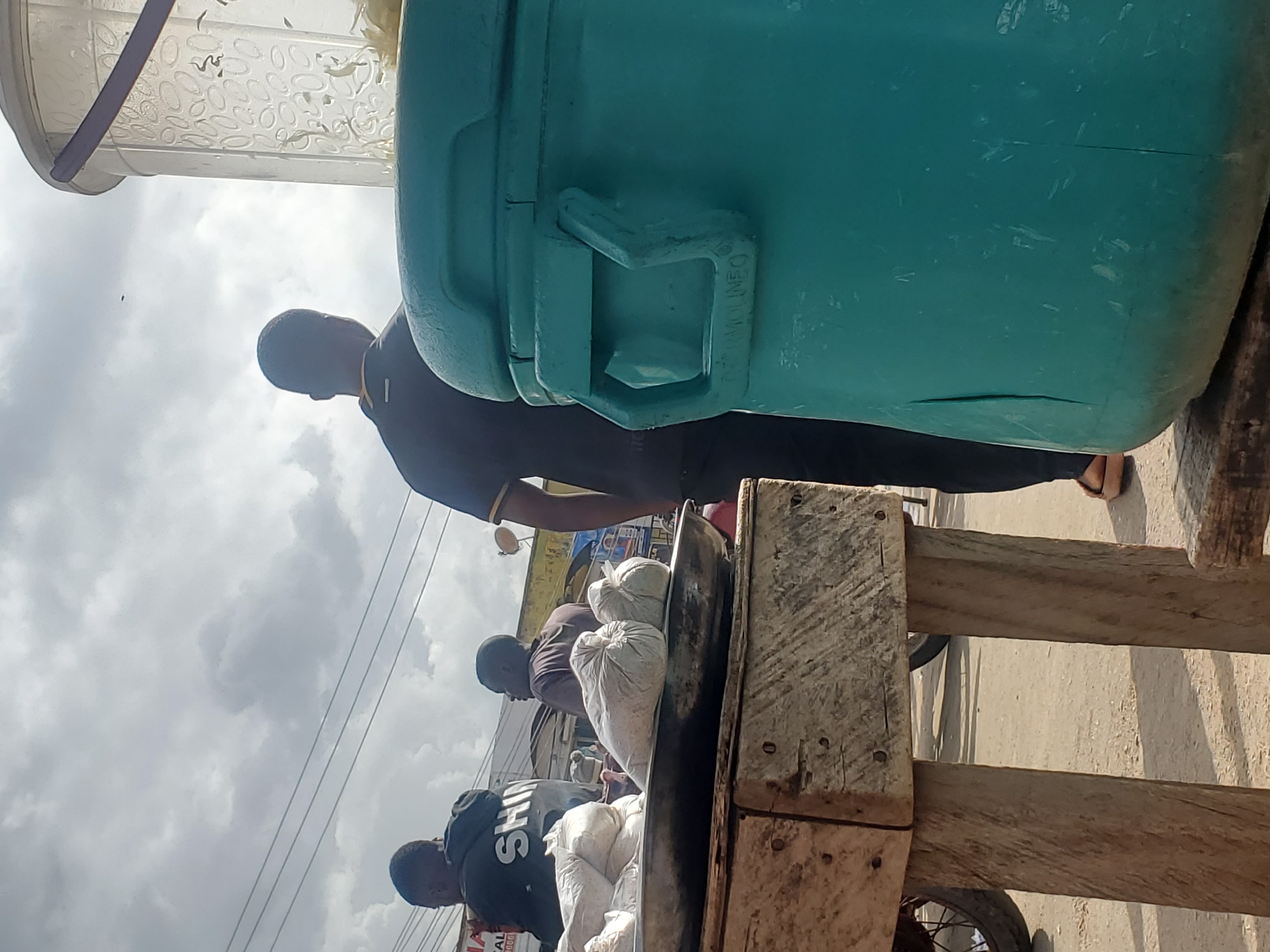 The image shows a street scene in Gwong, Nigeria, with people and goods displayed on a wooden cart. The sky is overcast, suggesting it might be daytime, possibly mid-morning or afternoon.
In the foreground, a man wearing a black t-shirt with "SH..." visible on the back is standing next to a cart loaded with sacks of what appear to be dried goods or grains. Behind him, another person in a dark t-shirt is also near the cart, with their face partially obscured. Further back, a man in a blue shirt is leaning forward, looking towards the cart.
The cart itself is made of rough, weathered wood, with a metal rim visible on a wheel at the bottom right. On top of the cart, several white sacks are piled up. There is also a large, teal-colored container visible in the upper right portion of the image, with a lid that has a patterned texture.
The scene appears to be a marketplace or a roadside stall, typical of many locations in Nigeria where vendors sell goods directly from carts or stalls. The overall impression is one of everyday commerce and activity. The people's postures suggest they are engaged in selling or attending to the goods. There are no strong indicators of specific emotions, but the scene is active.