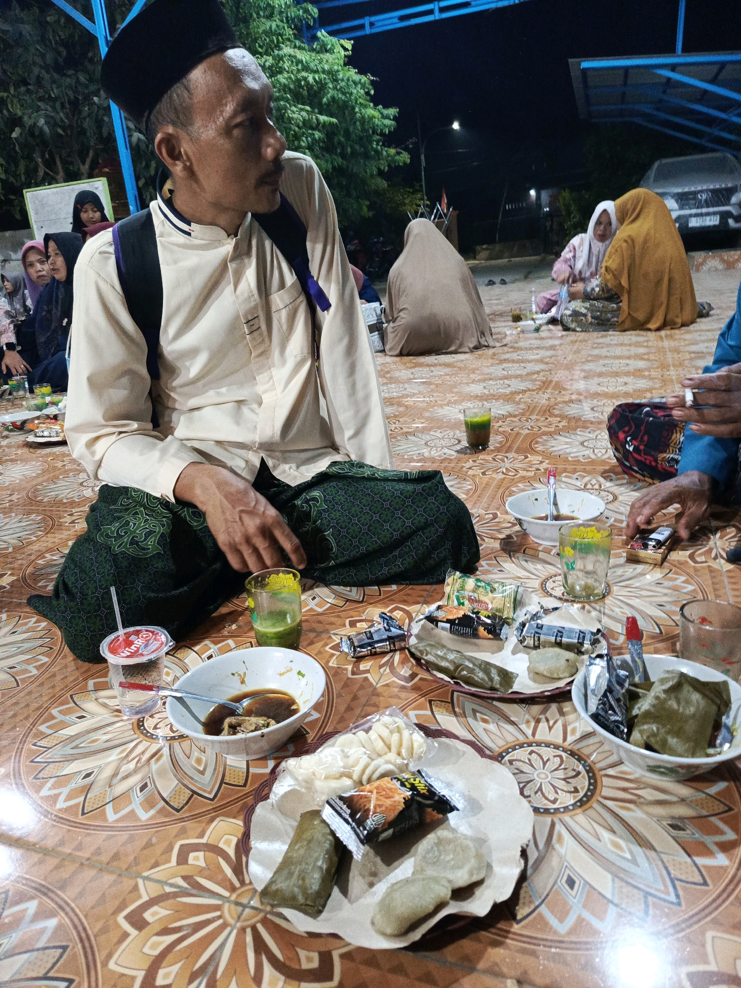 In Megang Sakti V, Indonesia, at night, several people are gathered on the floor, which is covered with a patterned rug. A man in the foreground, wearing a black peci hat and a light-colored, long-sleeved shirt, is seated. He has a green patterned sarong around his waist and a backpack slung over his shoulder. His right hand rests on his lap, while he looks to his right. In front of him, the rug displays various food items including bowls of soup and what appears to be some sort of wrapped food, along with glasses of green liquid, a plastic cup with a straw, and packets of snacks. In the background, other individuals are also seated on the floor, some wearing hijabs. One person is holding a cigarette and a lighter. The scene suggests a casual gathering or meal taking place outdoors under artificial lighting, with a car visible in the distance.
