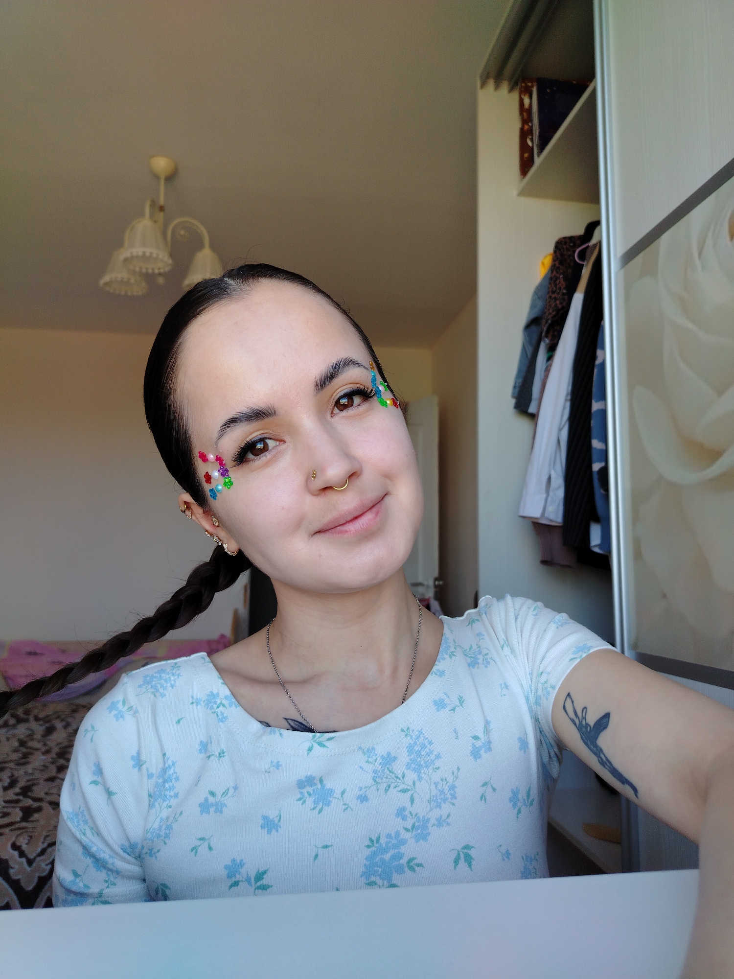 A medium shot photograph shows a woman with her hair pulled back into a single braid. Her left profile is towards the camera. The woman has brown eyes and fair skin. On her left temple, she has an arrangement of colorful floral and pearl-like embellishments. She also has a gold hoop nose ring and multiple gold earrings in her left ear. She wears a light blue floral patterned shirt and a silver chain necklace. On her right arm, she has a dark blue tattoo. The background features a closet with sliding doors, one of which has a floral design. A chandelier is visible on the ceiling. The location is Noviy, Russia.