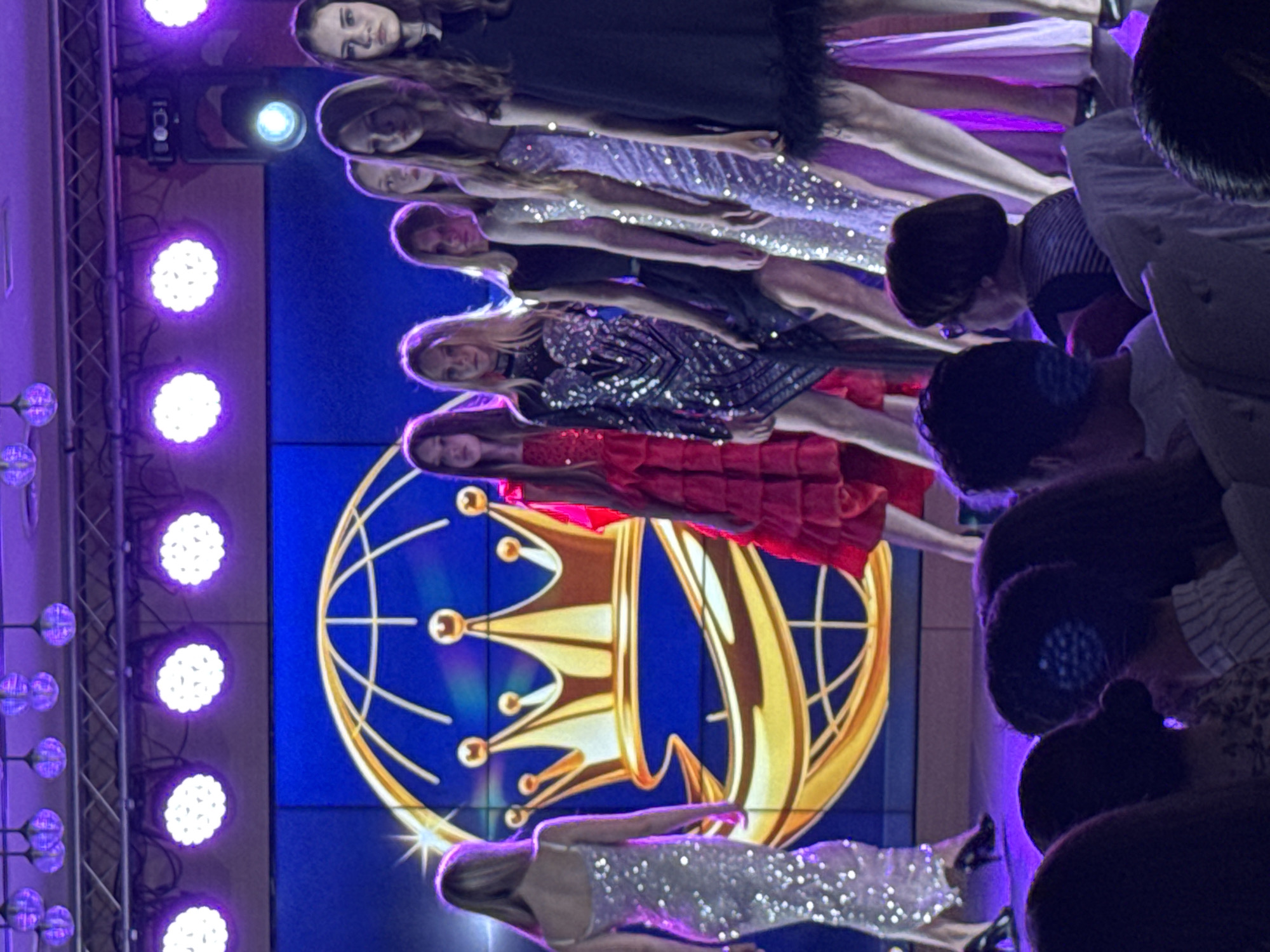 The image shows a group of people, mostly women, standing on a stage in what appears to be a fashion show or similar event. The stage backdrop features a large screen displaying a logo with a crown and globe design in gold against a blue background. To the left of the stage, there is a lighting rig with multiple purple spotlights. The environment suggests a venue in the city of Batumi, Georgia.