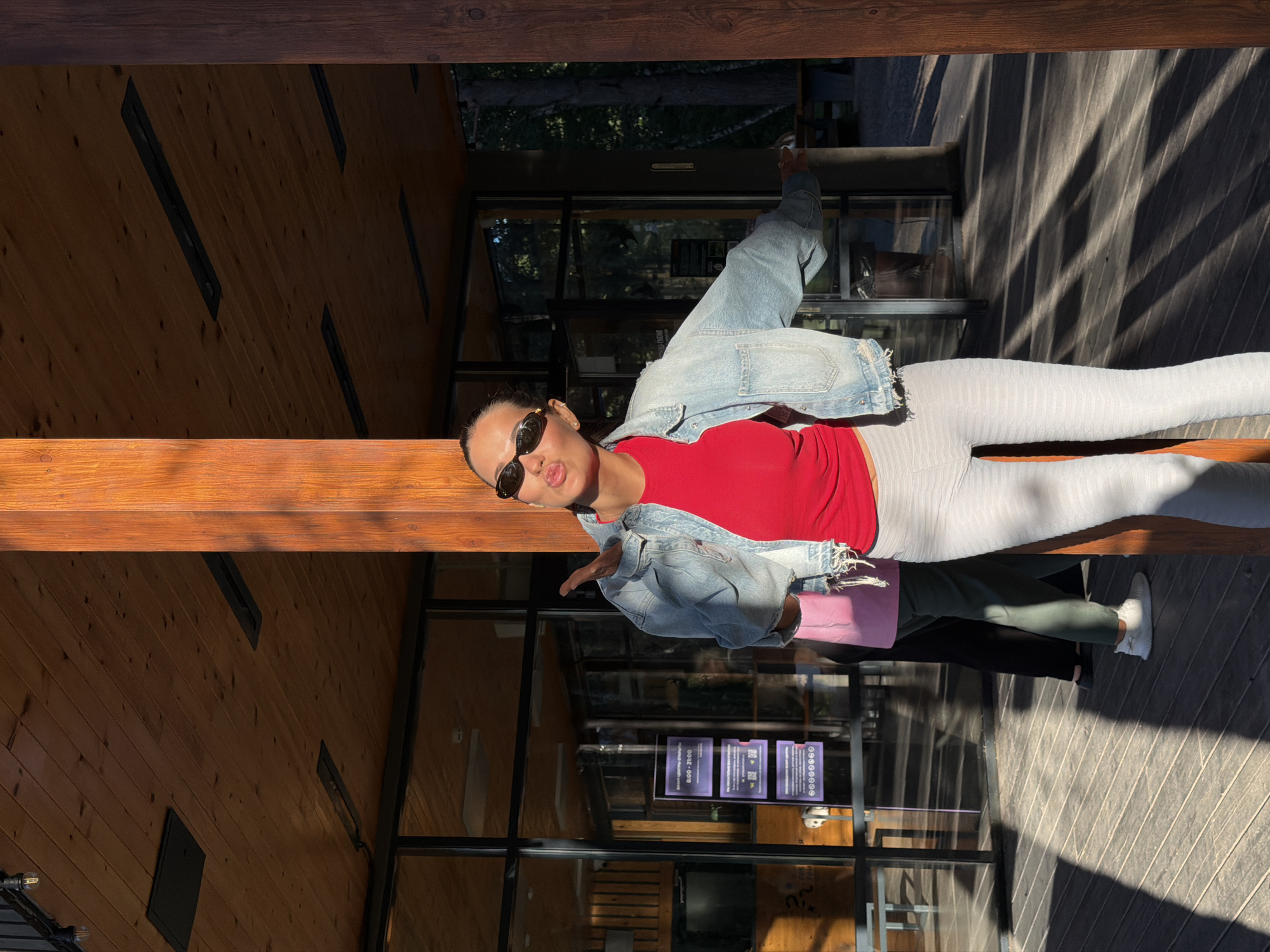 A young woman, wearing a red top, denim jacket, and light-colored leggings, stands on a wooden deck with a playful expression, blowing a kiss. She is partially framed by wooden pillars and a wooden ceiling with exposed beams and modern light fixtures. Behind her, large windows reveal a glimpse of trees and a building interior. Another person, wearing a pink shirt and dark pants, stands in the background. The scene is bathed in the warm, dappled sunlight of late afternoon, casting distinct shadows across the deck. The location is an outdoor area of a modern building, possibly a cafe or community center, in a park-like setting in Krasnoyarsk, Russia.