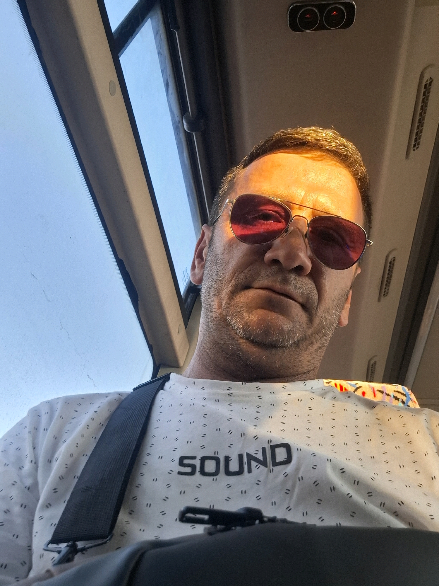 The image is a low-angle, selfie-style photograph taken from inside a vehicle, possibly a bus, as indicated by the windows and ceiling fixtures. The subject is a middle-aged man with fair skin, wearing sunglasses with pink lenses and a white t-shirt with a repeating small dash pattern and the word "SOUND" printed on it in black lettering. He has short, styled brown hair and stubble. The lighting suggests it is either early morning or late afternoon, with warm light casting shadows on his face. The background shows a bright sky through the vehicle's windows, and the interior of the vehicle features a beige-colored ceiling with air vents and small lights. No specific landmarks or indications place this image within Be'er Sheva, Israel.