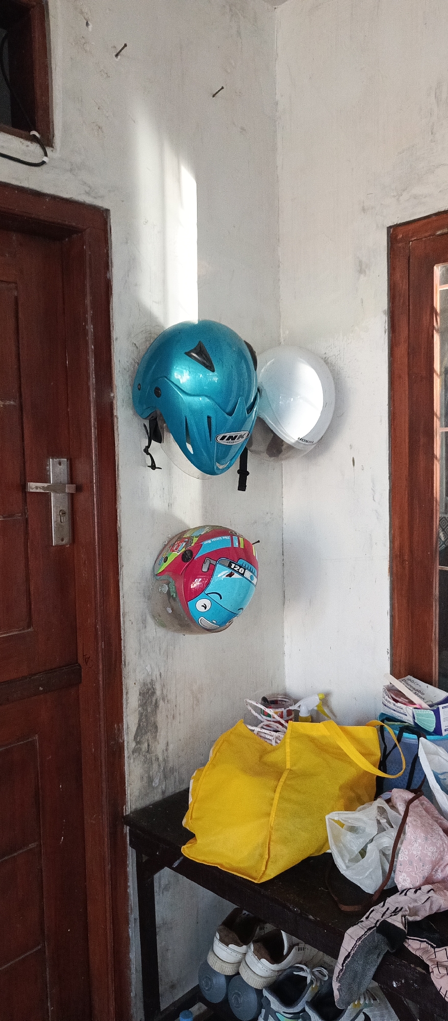 Two motorcycle helmets are hanging on a textured, white wall in a corner. The helmet on the left is bright turquoise with black accents and a clear visor. The helmet on the right is a solid white, also with a clear visor. Below the helmets, a wooden shelf holds a bright yellow tote bag, some plastic bags, and various clothing items, including socks and what appears to be a pink patterned garment. On the lower shelf of the same unit are several pairs of shoes, including sneakers. To the left of the helmets is a dark brown wooden door with a silver handle. To the right of the helmets is a wooden window frame. The overall environment appears to be an indoor, cluttered space, possibly a storage area or entryway.
