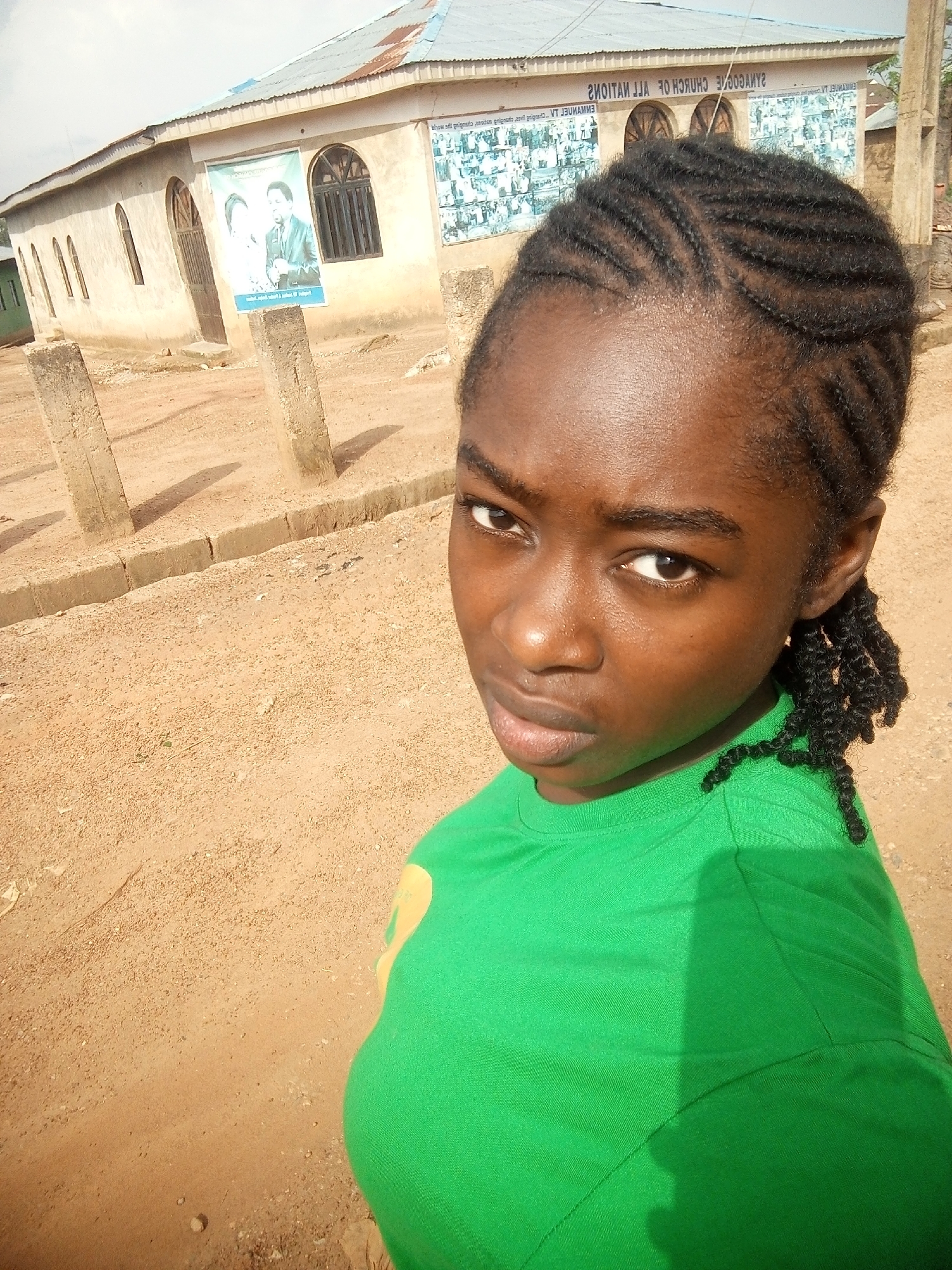 A young Nigerian girl with dark skin and braided hair poses for a selfie outside a building with a tin roof and arched windows. The building appears to be a church, as suggested by a poster on the exterior depicting a man and woman. The ground is covered in sand and loose gravel, and there are several concrete posts in the foreground, casting long shadows. The girl wears a bright green t-shirt. The scene is set in Rafin Par, Nigeria.