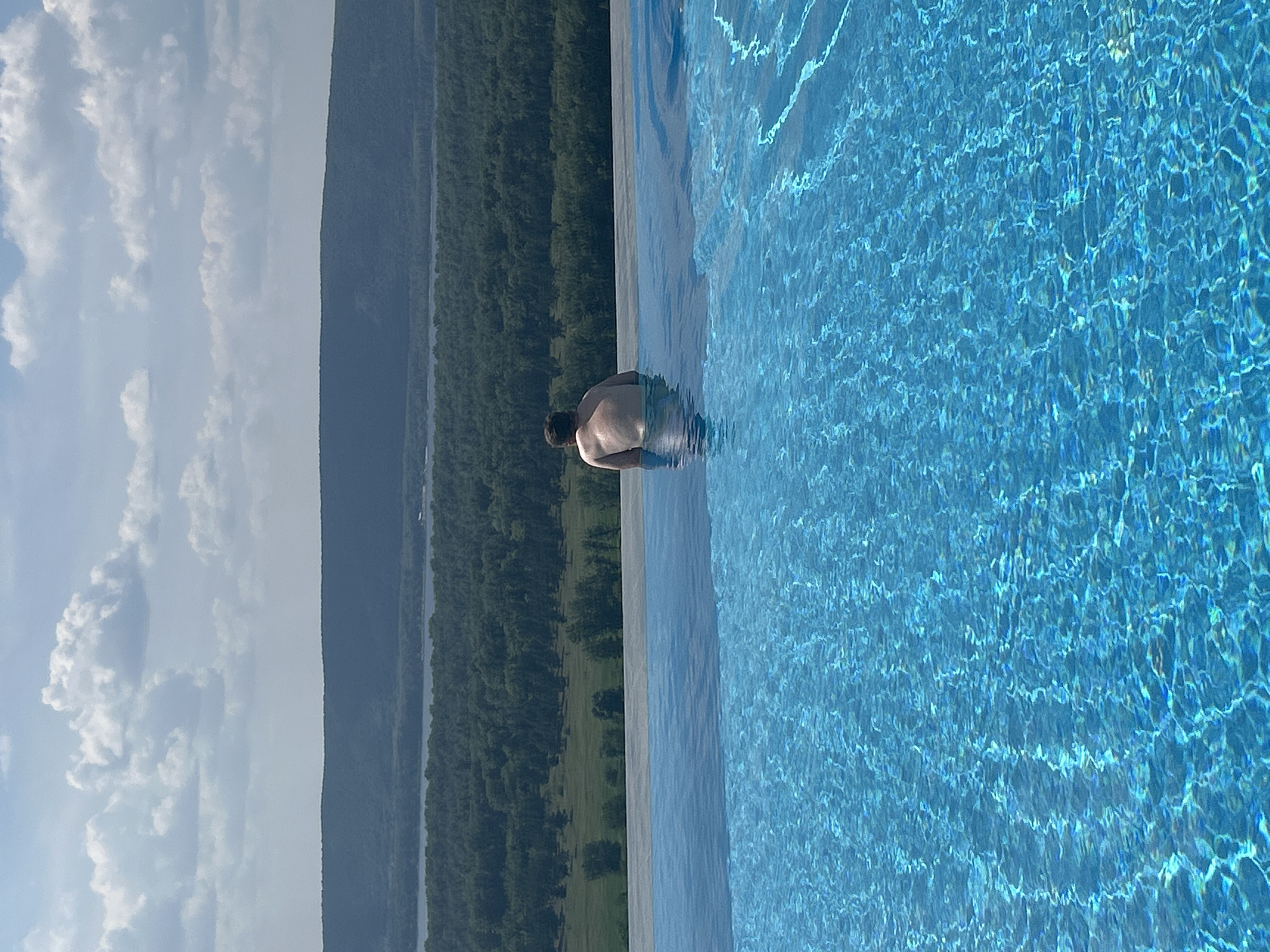 The image shows an infinity pool with bright blue water. In the pool, a person from behind is submerged up to their torso. They are facing away from the camera, looking out at a landscape of green trees and a body of water, possibly a river or lake, bordered by hills in the distance. The sky above is blue with scattered white clouds. This scene could be located in the vicinity of Novokuybyshevsk, Russia, a city known for its surrounding natural beauty.
