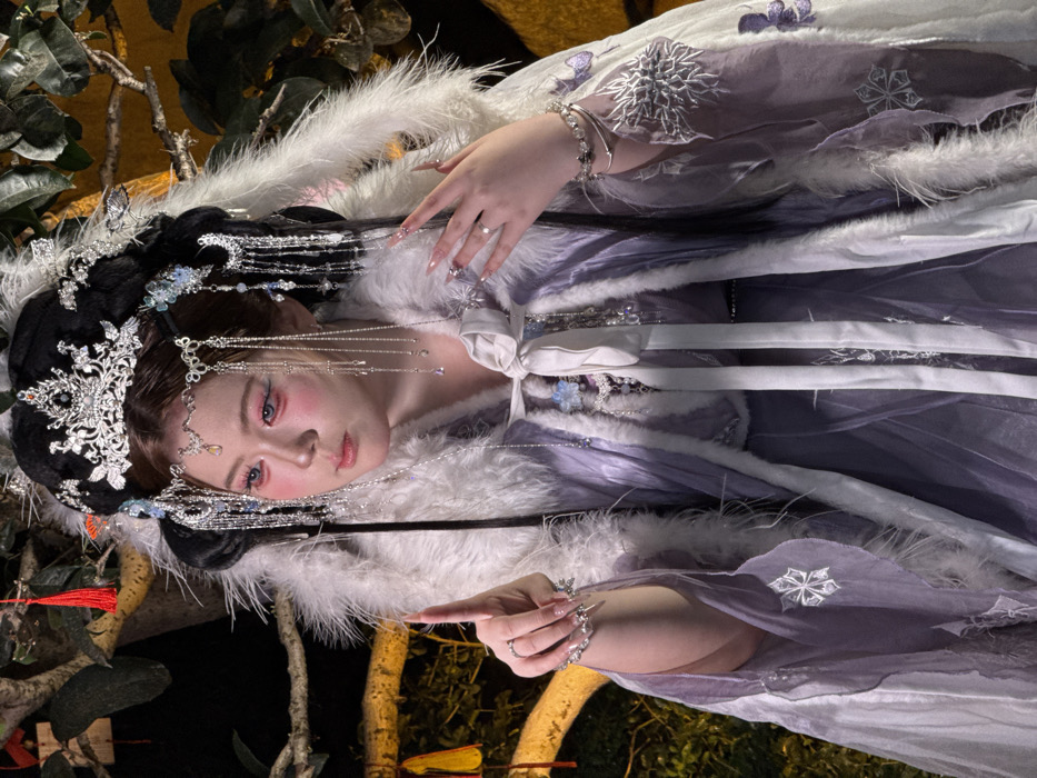 A female individual is depicted in an elaborate, multi-layered historical or fantasy-style Chinese costume, set against a dimly lit, treed background. She has fair skin, prominent pink blush under her eyes, and light blue contact lenses. Her elaborate black wig is styled high and adorned with a large, ornate silver-colored crown, intricate filigree, and numerous dangling silver chains with pendants. She wears a white cape trimmed with thick white faux fur, featuring silver snowflake-like embroidery and purple butterfly appliqués. Beneath the cape, a purple garment is visible, tied at the chest with a white ribbon. Both hands are adorned with silver-colored rings and elongated, pointed decorative nail extensions. Her right arm is raised with the index finger pointing upwards and slightly forward. Her left hand rests near her chest. The background features dark foliage, a faint yellow light source, and a hanging red tassel ornament. The scene is located in Yuzhong District, China.