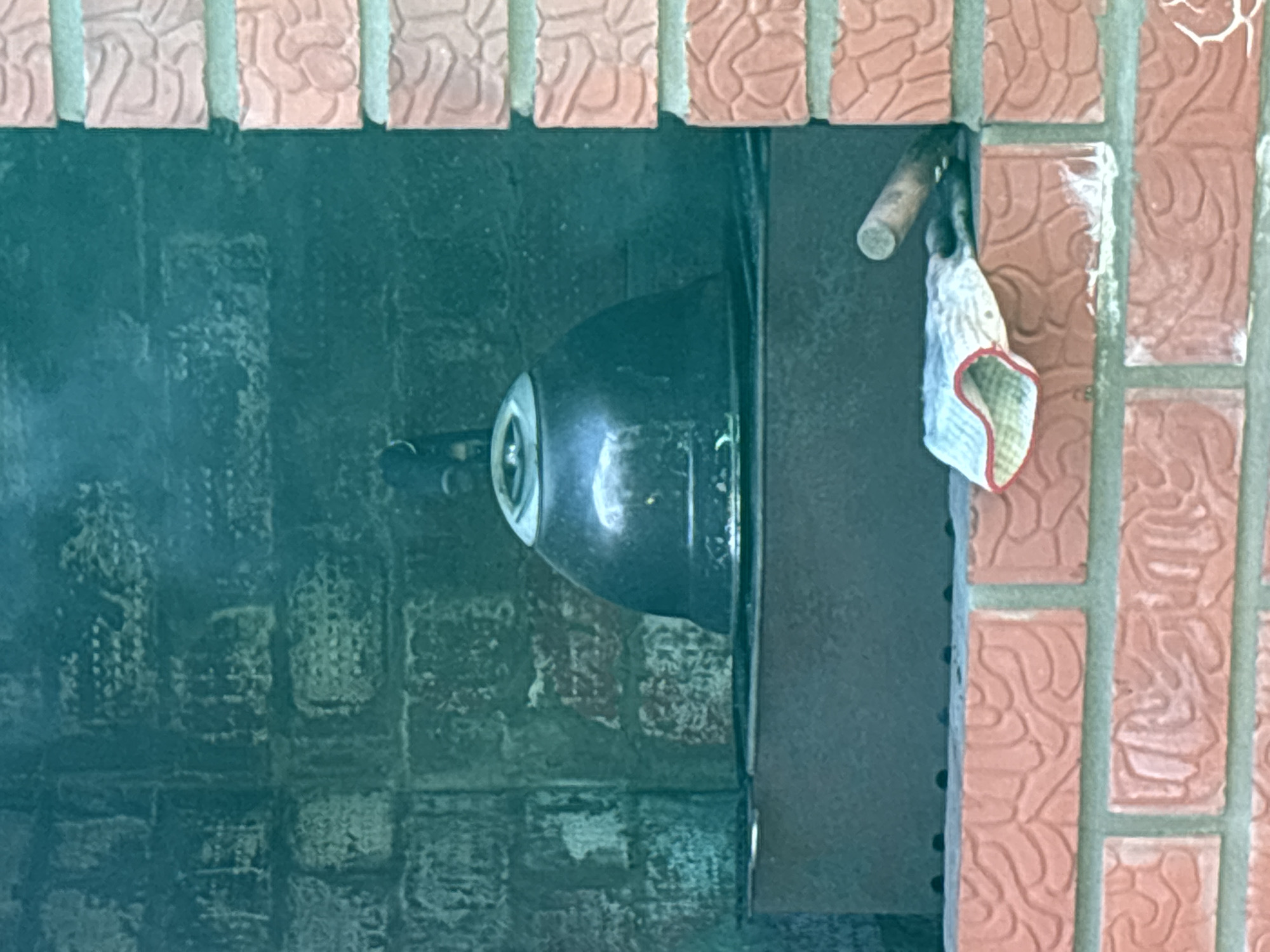 The image depicts a barbecue pit constructed of patterned red bricks in Chaikovsky, Russia. A metal grill is positioned inside the pit, with a blackened dome covering the cooking surface. Hanging on the right side of the grill, outside of the fire pit, is a worn rag with a red trim. The interior brickwork of the pit appears darkened with soot.