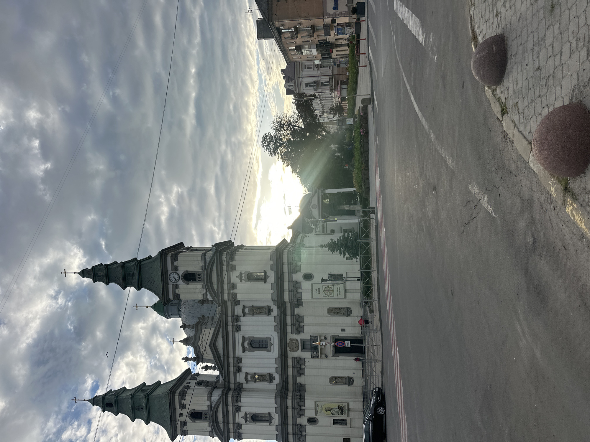 The image shows the Ternopil City Council building and the Ternopil Regional Academic Ukrainian Drama Theatre, located in Ternopil, Ukraine. The perspective is from the street level, looking towards the buildings. The sky is overcast with sunlight breaking through the clouds, casting rays of light. The foreground features an asphalt road with pedestrian crossings and some concrete bollards. In the midground, the impressive architecture of the city council building is visible, with its distinctive towers and ornate facade. A few people are seen near the entrance of the building. To the right of the council building, the drama theatre is partially visible, along with adjacent urban buildings. There is a tree and some greenery in front of the theatre. The overall scene suggests an urban setting with a mix of historical and modern architecture under a dynamic sky.