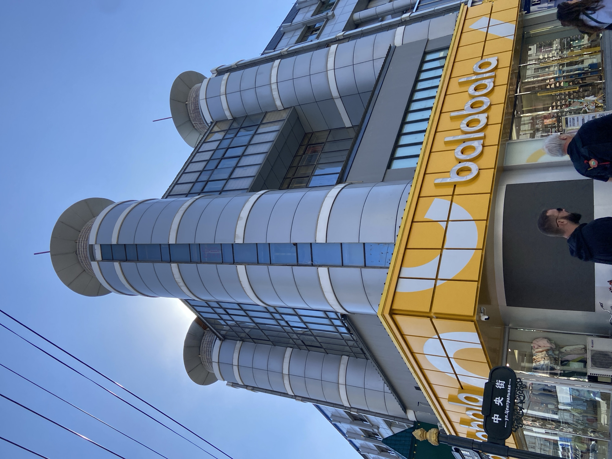 The image shows a street view of a modern building in 兴安街道, China. The building has a distinctive design with cylindrical towers and large glass windows. The ground floor features a bright yellow storefront with the logo "balabala" prominently displayed, suggesting it is a retail establishment, possibly for children's clothing given the brand. A street sign in Chinese characters and Cyrillic script indicates the location "ул. Центральная" which translates to "Central Street". Two men are partially visible in the foreground, walking away from the camera, their backs to the viewer. The sky is clear and blue, with some sunlight glinting off the building's facade. Electrical wires are visible in the upper portion of the image.