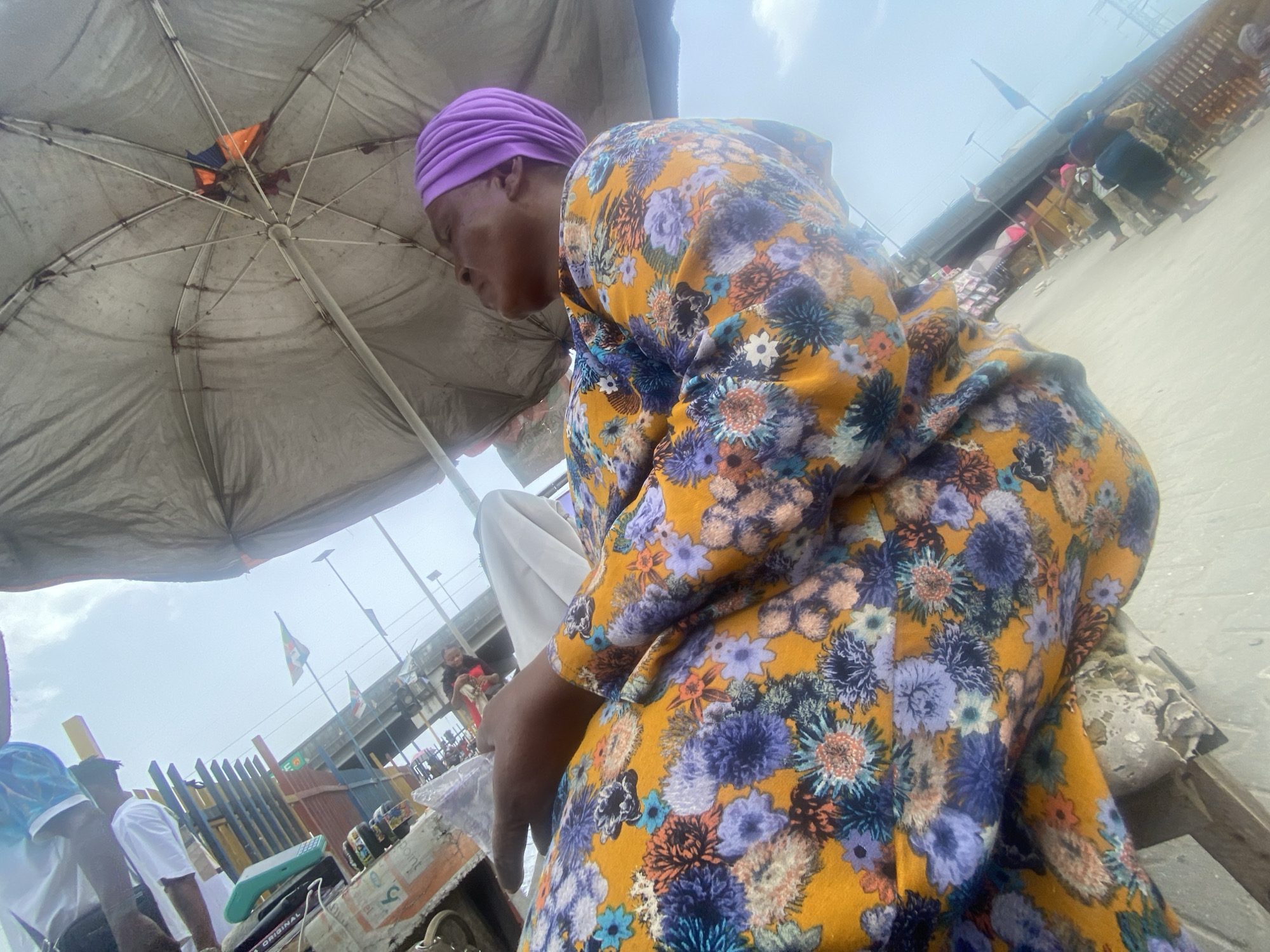 A woman wearing a purple head covering and a yellow and blue floral patterned dress sits under a large, weathered gray umbrella. The scene appears to be in a busy outdoor market or commercial area in Lagos, Nigeria. In the background, there are other people, structures including a bridge or overpass, and flags suggesting an urban environment. The camera angle is low, looking upward towards the woman and the umbrella.