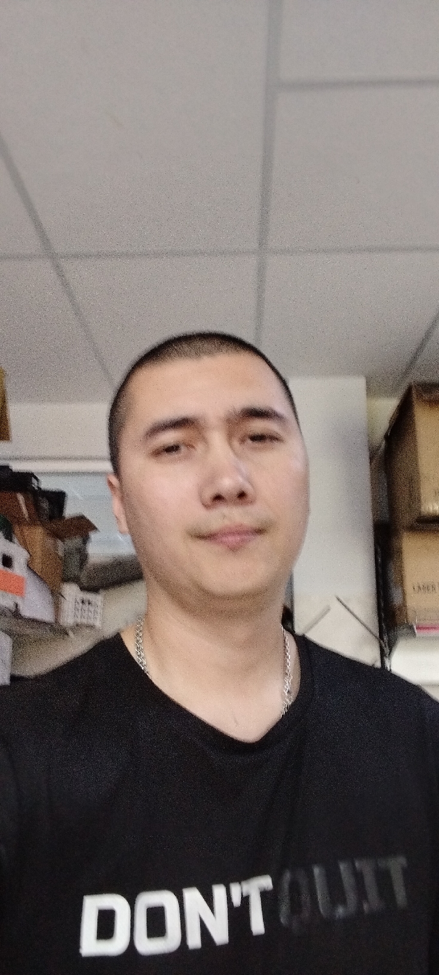 The image shows a head-and-shoulders shot of a man with a shaved head and a silver chain necklace. He is wearing a black t-shirt with the words "DON'T QUIT" printed on it in white lettering. The man is facing the camera directly, and the background is blurred, showing a ceiling with fluorescent lighting and some boxes stacked behind him. The overall scene appears to be an indoor environment, possibly an office or storage area. There is no indication of the city of Aktobe, Kazakhstan in the image.