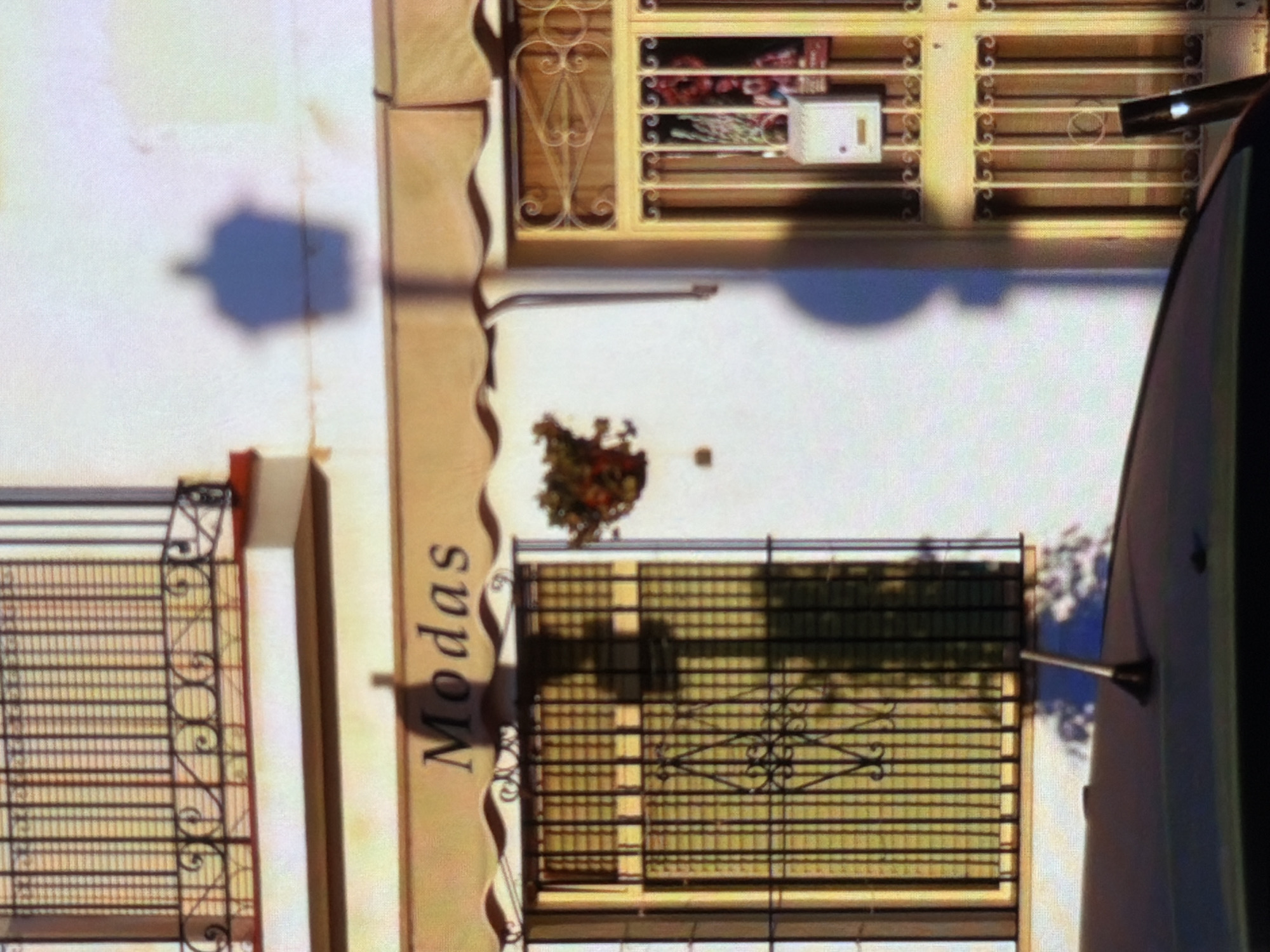The image shows the facade of a building with a storefront. The storefront has a beige awning with the word "Modas" printed on it. Below the awning, there is a window with dark metal bars and green blinds. To the right of the window, there is a yellow-painted door with decorative metalwork and a small white mailbox. A hanging planter with red and green foliage is visible on the wall to the left of the door. Shadows of objects, possibly people, are cast on the white wall. A car antenna is visible in the foreground on top of a dark car. The building appears to be in an urban or suburban setting.