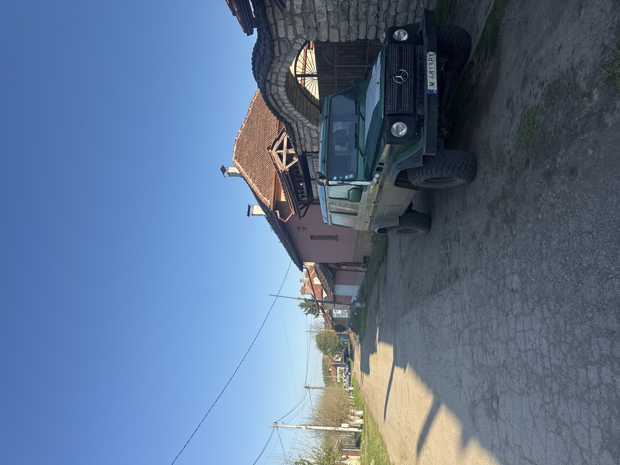 A green Mercedes-Benz G-Class (license plate M 6813 BX) is parked on the side of a narrow road in what appears to be a village. The vehicle is parked next to a stone archway that leads to a building. Other buildings and houses are visible along the road in the background, along with utility poles and vegetation. The sky is clear and blue. Based on the provided location (Lehchevo, Bulgaria), the buildings' architecture aligns with typical village structures in that region.