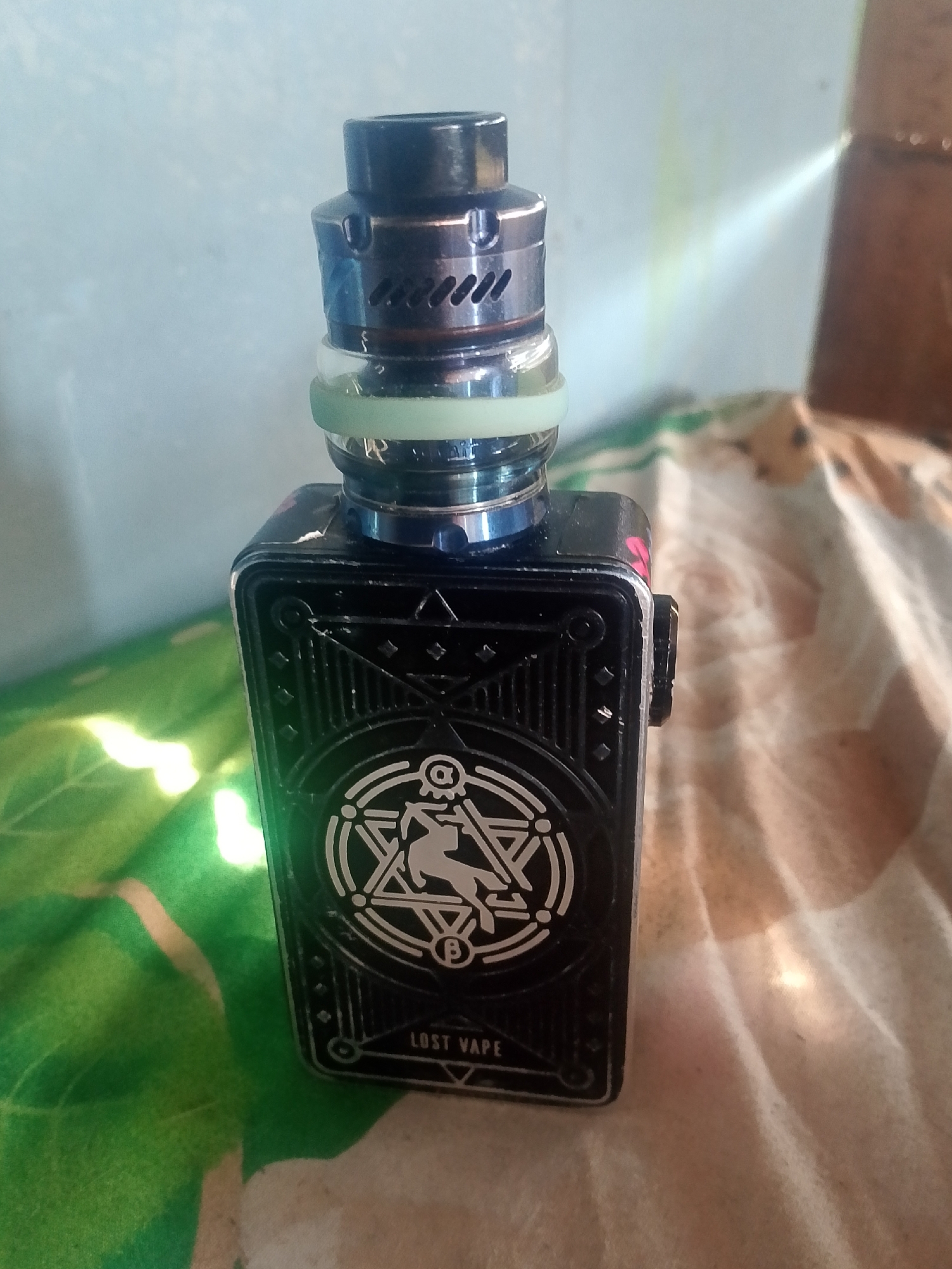 A black vape mod with a stylized symbol on its side is positioned on a surface that appears to be a patterned cloth, possibly a bedspread. Above the vape mod sits a metallic atomizer with visible air vents. A light green o-ring is present between the mod and the atomizer. The background is a light-colored wall with a faint yellow-green streak, suggesting a light source or reflection. The overall scene is indoors. The location is identified as Tatah Bangkal, Indonesia.