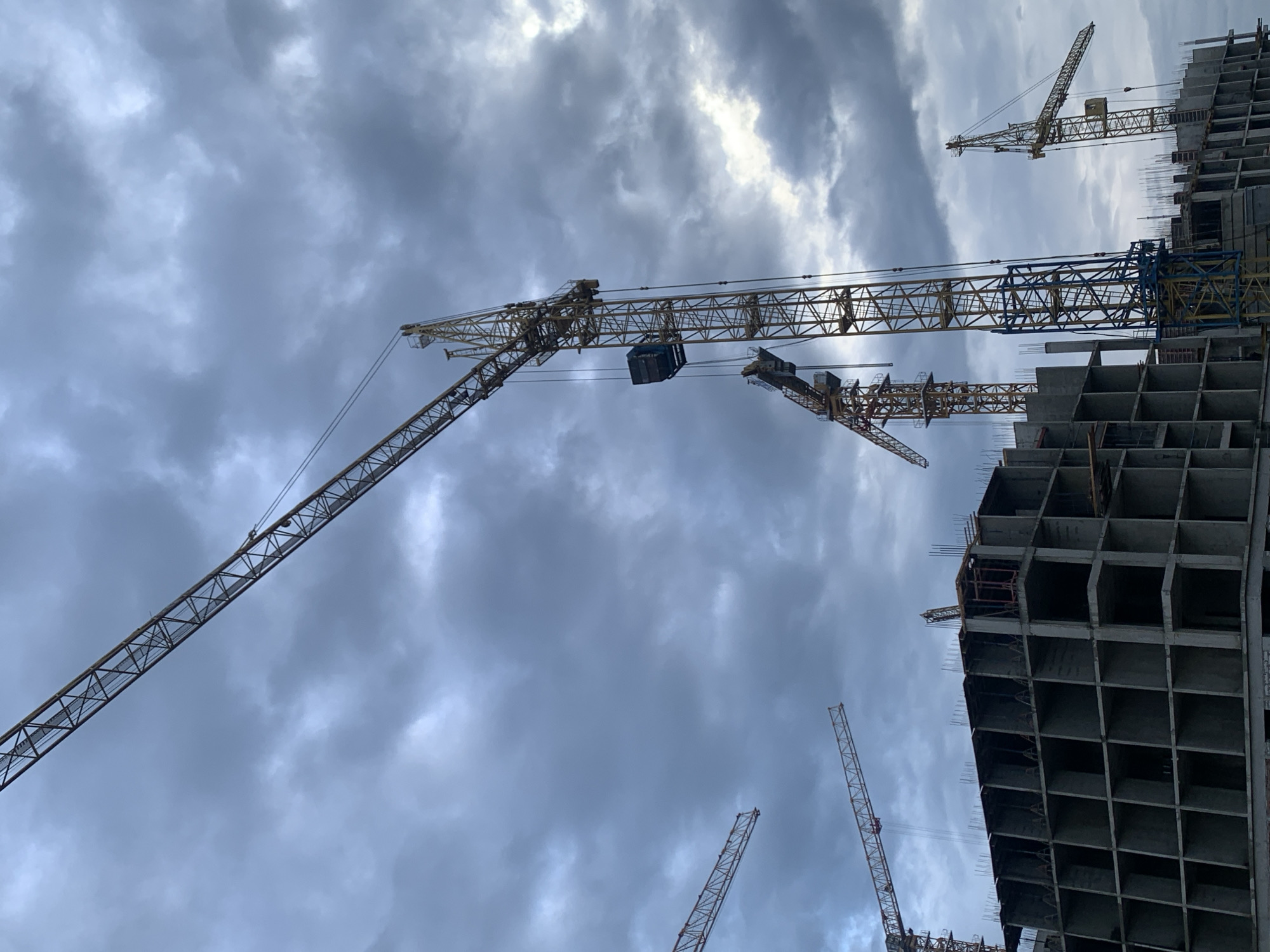 The image depicts a construction site in Stavropol, Russia, under a cloudy sky. Multiple tall yellow construction cranes are visible, with one prominently positioned in the foreground. A large, unfinished concrete building with a grid-like structure occupies the lower portion of the frame. The building's floors are open, and rebar protrudes from the top, indicating ongoing construction. The overall scene is one of industrial activity and development in an urban setting.