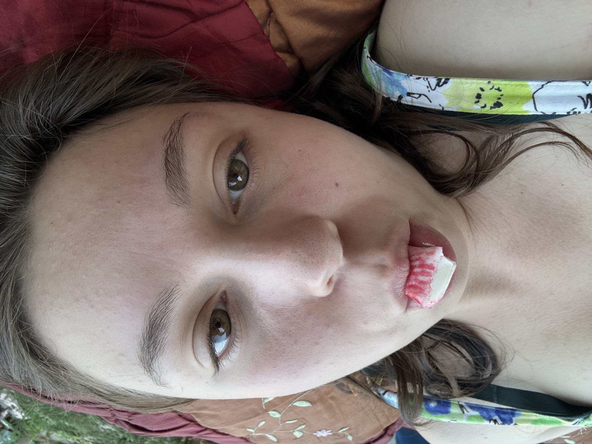 The image is a close-up, overhead shot of a young person lying down, with their head tilted back. They have fair skin, brown eyes, and dark brown hair that falls around their face and shoulders. A colorful, floral-patterned strap, possibly from a piece of clothing, is visible across their chest.
Their face is the primary focus. A piece of something white and red, possibly food or a bandage, is visible on their lips. Their expression is neutral or slightly relaxed. The background consists of textured fabric, one piece of which is a deep red, and another a muted brown with embroidered floral details.
The image appears to have been taken outdoors, suggested by the natural lighting and the hint of greenery in the bottom left corner. The time of day is likely daytime, given the brightness. No visible text is present in the image. The location context is Shymkent, Kazakhstan.