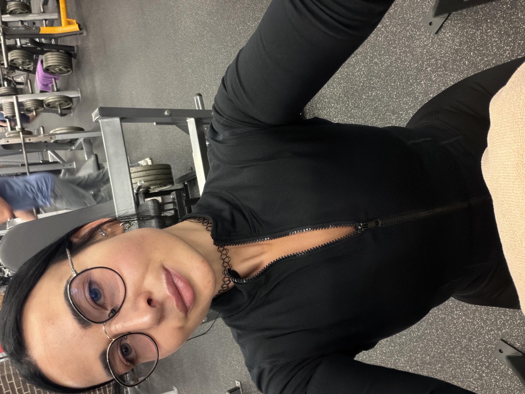 The image depicts a female individual taking a selfie in an indoor gymnasium, potentially located in Saint Petersburg, Russia. The woman, who appears to be in her late 20s to mid-30s, occupies the foreground. She has dark hair, a light complexion, and wears round glasses with dark frames and gold-colored accents, behind which her eyes appear light blue or grey. Her lips are full and have a pink tint. She is wearing a form-fitting black long-sleeved athletic top featuring a silver-colored zipper, which is partially unzipped to reveal a small portion of her upper chest. A black, patterned choker necklace is worn around her neck. Multiple small silver earrings are visible in her left ear. She is looking directly at the camera with a neutral to slight smile. A light-colored, textured towel, possibly beige, is visible in the bottom right of the frame, partially obscuring the equipment she is likely seated upon.
The background shows a gym environment with speckled dark grey rubberized flooring. Various pieces of gym equipment are visible, including metal barbell racks heavily loaded with weight plates, weight benches (one of which has a purple towel draped over it), and other indistinct metal exercise structures. Two blurred male figures are present in the mid-ground. The figure on the left is partially visible, wearing a blue shirt and grey pants, appearing to be walking or standing near equipment. Another less distinct male figure in a blue top is visible further right, also within the gym setting. All background elements are out of focus, consistent with a selfie perspective.