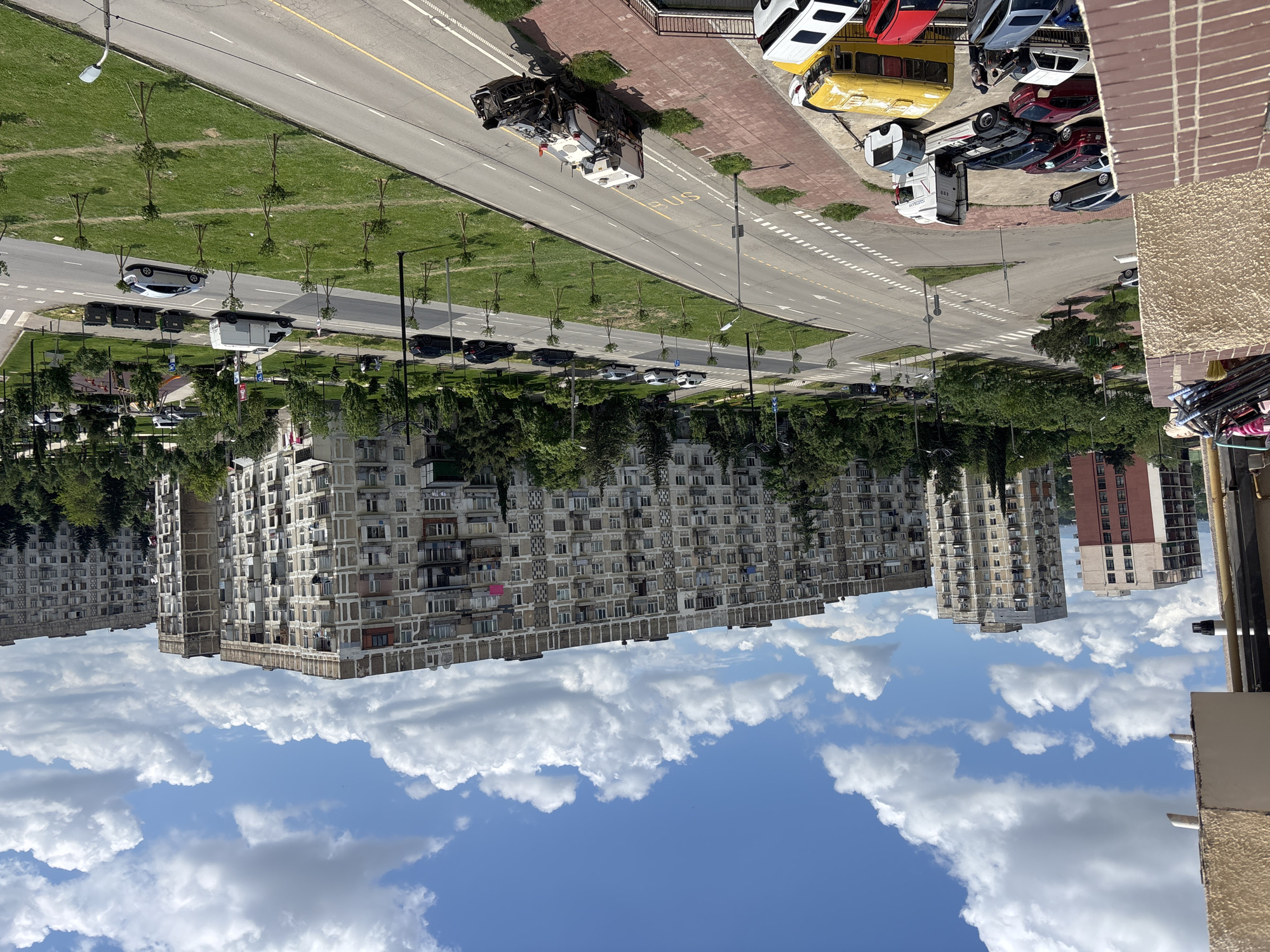 An elevated, slightly wide-angle shot shows a cityscape dominated by Soviet-era apartment buildings under a partly cloudy sky, likely taken in Tbilisi, Georgia. The focus is on a main road with sparse traffic. On the right edge of the road, a cluster of parked vehicles includes cars, a yellow bus, and a truck. A well-maintained median divides the road, featuring grass and young trees. High-rise residential buildings with a repetitive, rectangular design fill the horizon. In the foreground, a balcony with household objects provides perspective. The sky contains scattered white clouds against a bright blue background, adding to the depth of the scene.