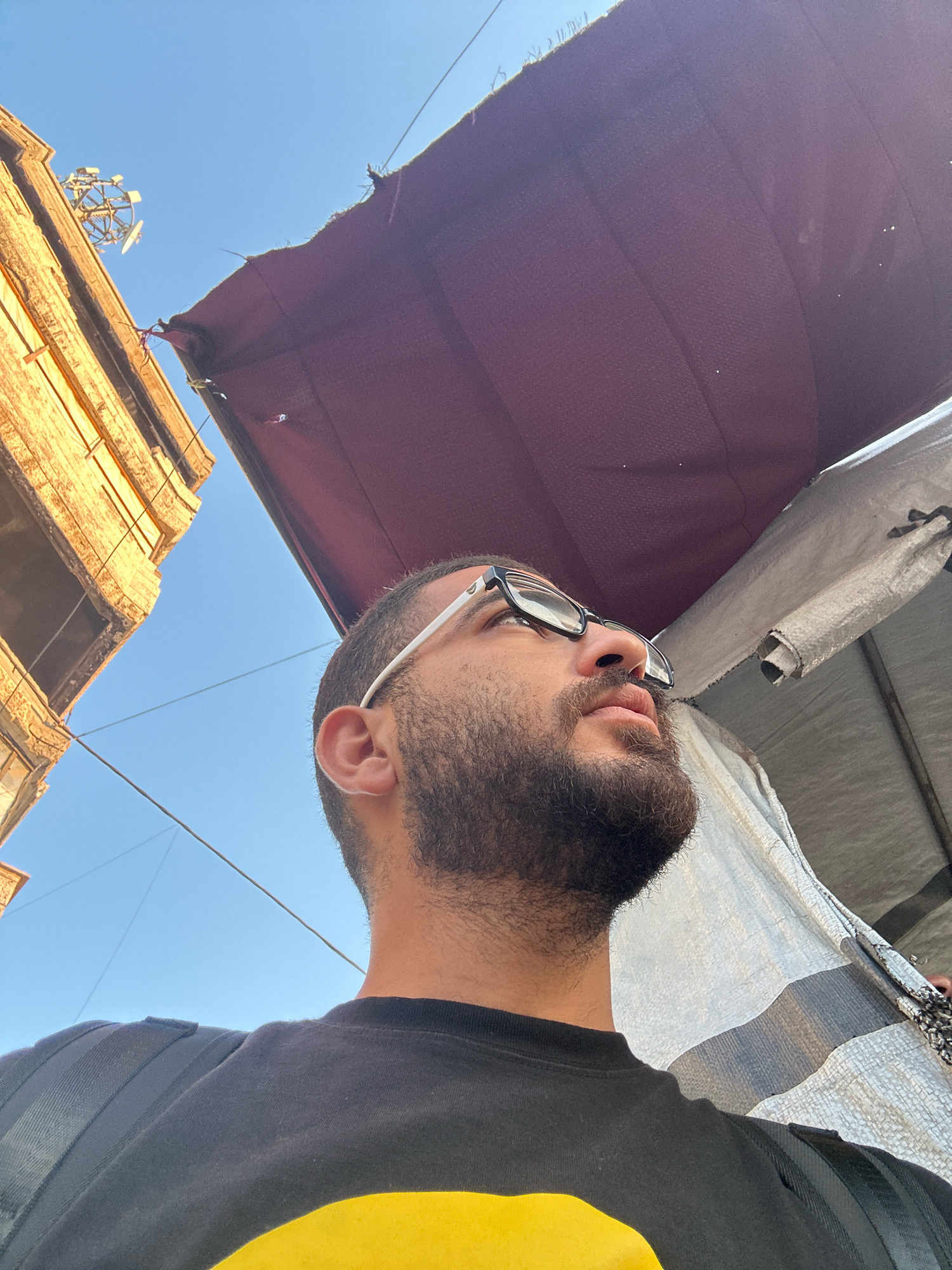 A low-angle, close-up shot shows a man from the chest up, looking upwards and to the right. He has dark, short hair, a full beard, and is wearing black-rimmed glasses and a black t-shirt with a bright yellow graphic design. His ear is visible on the left side of the frame. Behind him, a maroon tarp or awning stretches across the upper portion of the image, casting a shadow. To the left of the tarp and behind the man, an old, tan-colored building with visible architectural details and some disrepair is present. In the distance, against a clear blue sky, a cell phone tower is visible atop a structure. A white, textured tarp or canopy is visible to the right of the maroon tarp, partially obscuring the background. The setting appears to be an urban environment.