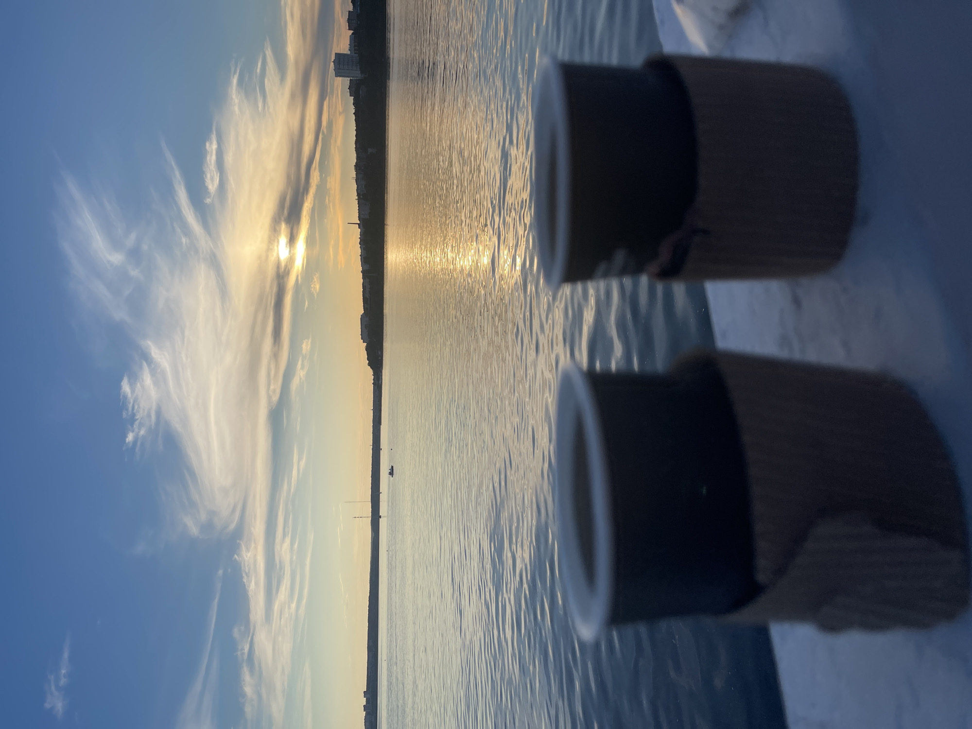 Two dark cups with brown cardboard sleeves are in the foreground, out of focus, on a white surface. In the background, a body of water reflects the setting sun, which is partially obscured by clouds. A city skyline with buildings and a tall tower is visible on the horizon across the water. A sailboat and a small boat with people are in the distance on the water. The sky is blue with wispy white clouds.