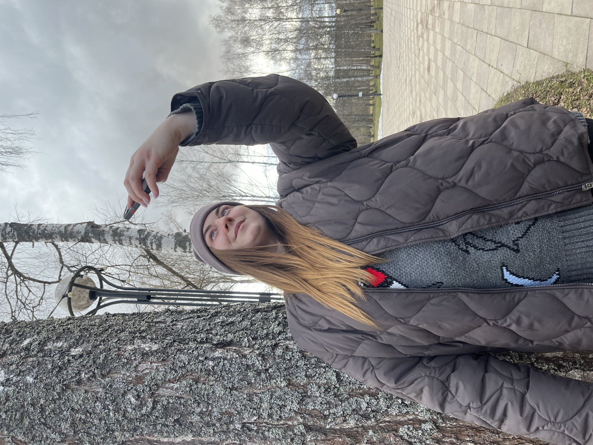 A young woman is holding a small black object, possibly a vape or lighter, near a tree in what appears to be a park or outdoor recreational area. She is wearing a gray knit hat, a brown quilted jacket, and a gray sweater with an animal design. The park is visible in the background with bare trees and a tiled walkway. The location is Vysokovsk, Russia.