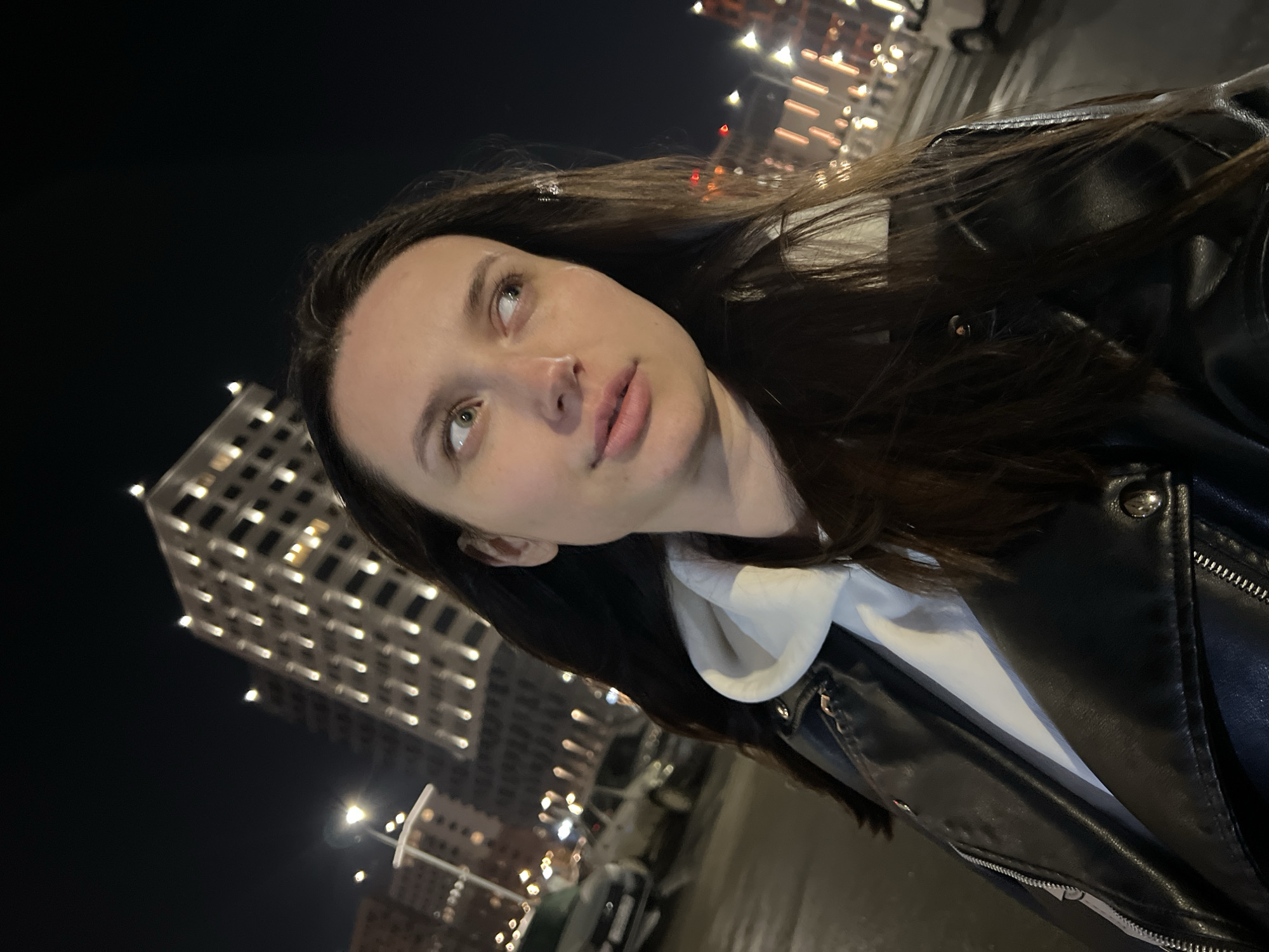 A woman with long brown hair stands in front of a building in Tyumen, Russia at night. She is wearing a black leather jacket over a white hoodie. The building behind her is lit up with many windows. Streetlights are visible in the background, along with cars. The woman is looking upwards with a neutral expression.