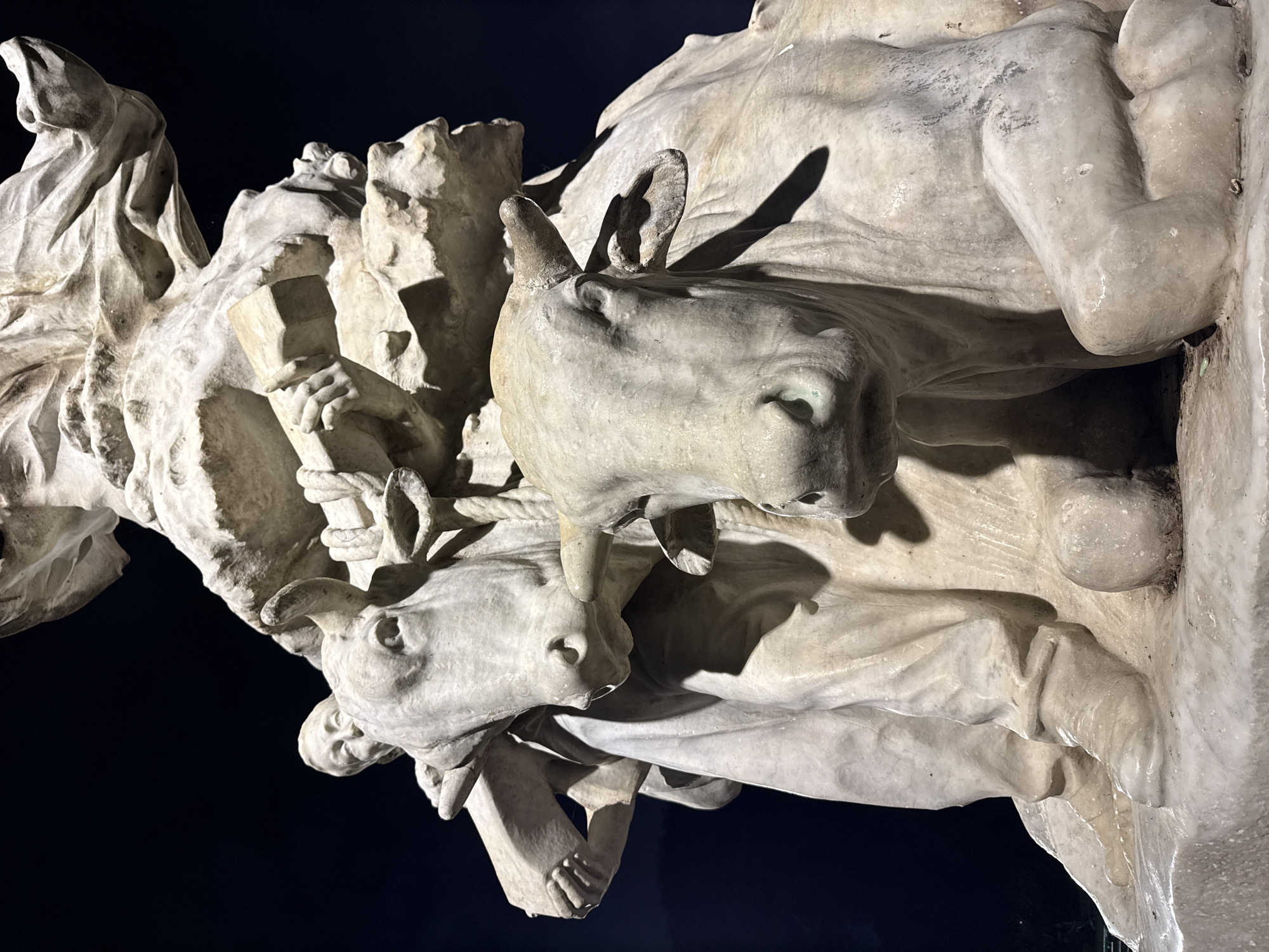 This media file features a detailed view of a large, complex marble sculpture, dramatically illuminated at night. The sculpture depicts a dynamic scene involving multiple figures, predominantly two donkeys or mules and several human figures.
The central focus is on two prominent animal heads, likely donkeys or mules, carved from light-colored stone. One animal faces left, its ears casting long shadows, while another is visible below and to the right, facing right. Both animals appear to be harnessed or tethered with ropes, suggesting a theme of labor, journey, or a procession. Associated with these animals are human figures, though only parts are clearly visible; a hand holding a block-like object (possibly a book or tool) is seen near the top left, and another human face is discernible looking downwards.
The sculpture is rendered in white or light grey stone, likely marble, and shows signs of weathering and some green/dark discoloration in crevices, indicating age or outdoor exposure. The deep, dark background signifies nighttime, with powerful artificial lighting casting stark contrasts and pronounced shadows that enhance the texture and three-dimensional quality of the carving. The overall composition is dynamic, possibly part of a larger monument. No text is visible within the image. The location context provided is Buenos Aires, Argentina.