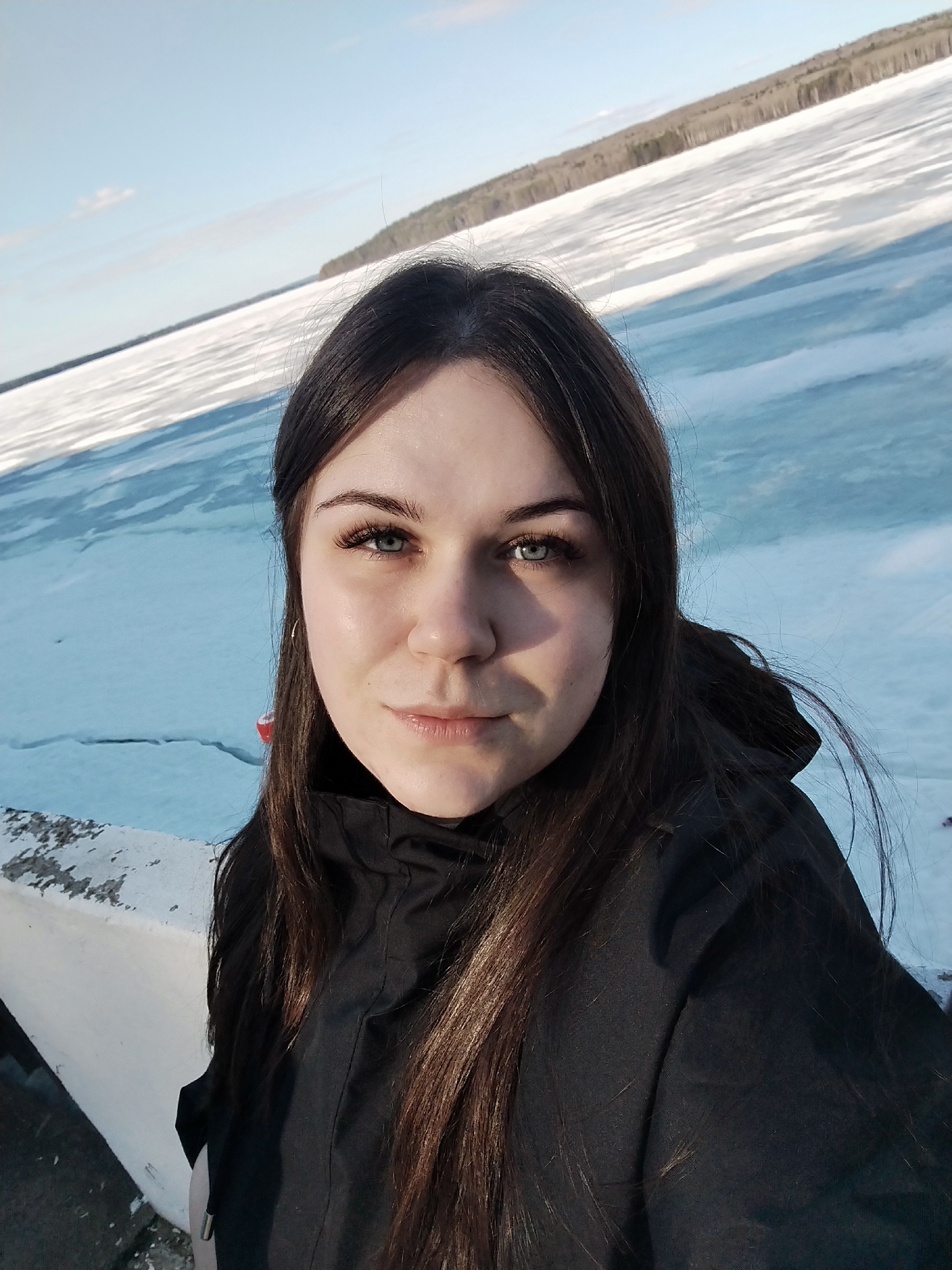 A woman with long, dark brown hair and light blue eyes stands in front of a frozen body of water, likely the Kama Reservoir, near Chaikovsky, Russia. She wears a black jacket. The sky is light blue. The water is frozen, with visible cracks. A treeline is visible in the background. A concrete barrier is in the foreground.