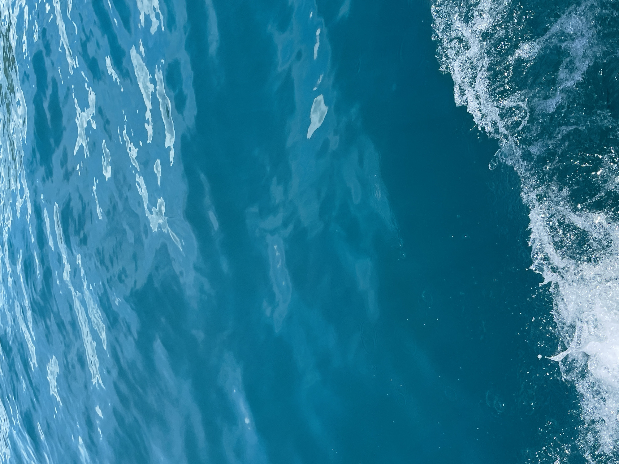 The image shows a section of ocean water. The water is primarily a deep turquoise color with rippling patterns across the surface. A foamy white wave crests along the right side of the frame, showing bubbling and splashing. The left side of the image reflects light in a manner that produces lighter shades. The specific location within Nha Trang Ward, Vietnam, cannot be discerned from the visual content alone.