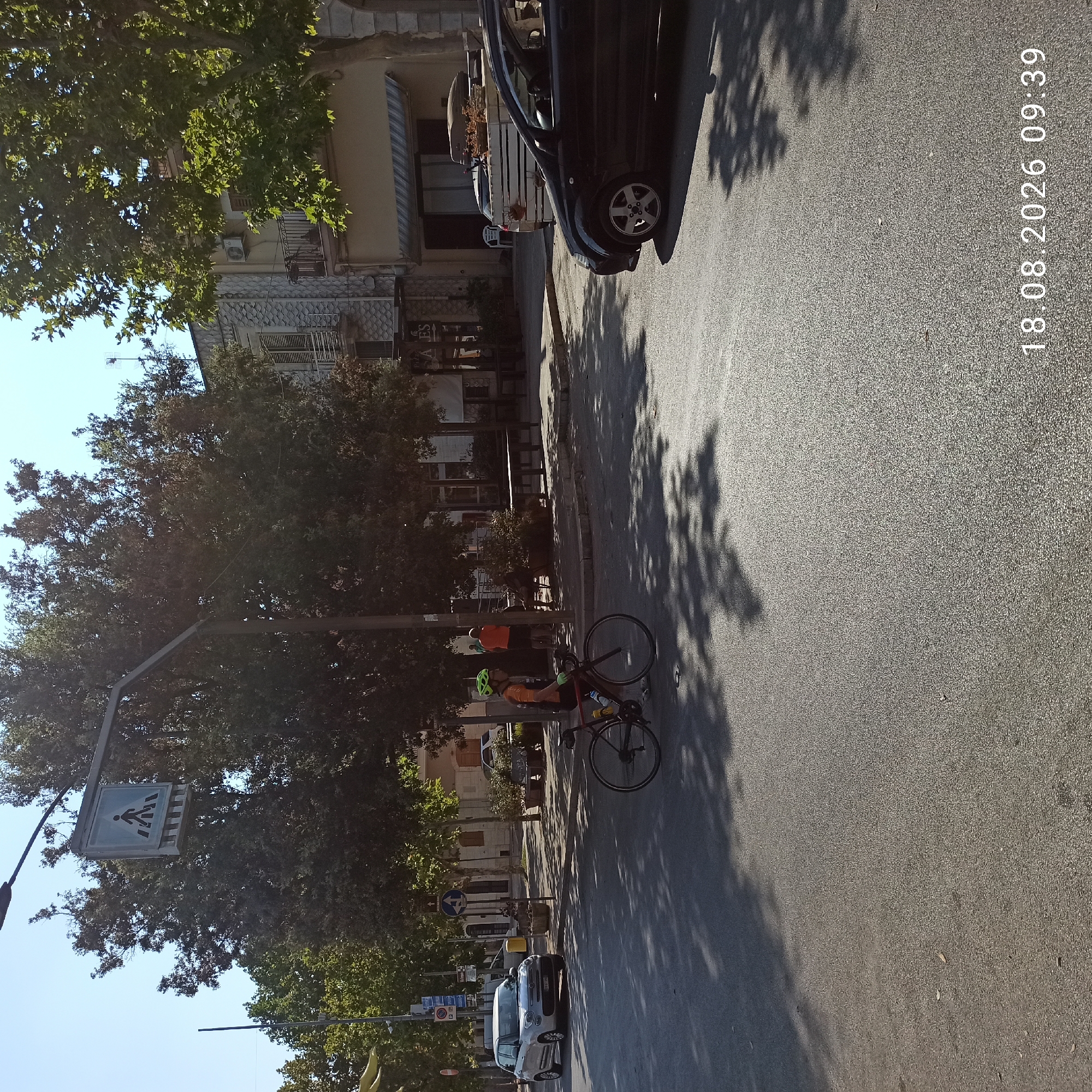 The image captures a street scene in Bitonto, Italy, on August 18, 2026, at 09:39. The foreground is dominated by an asphalt road with dappled shadows from trees. A cyclist wearing a green helmet and an orange and black jersey is in motion on the right side of the road, riding a bicycle. In the mid-ground, a light-colored car is parked on the left side of the street. Across the street, there are trees, buildings with balconies and shutters, and outdoor seating areas, suggesting a commercial or residential area. A pedestrian crossing sign is visible, mounted on a pole. In the background, more trees and buildings are present under a clear blue sky. The overall scene depicts a sunny day in a town.