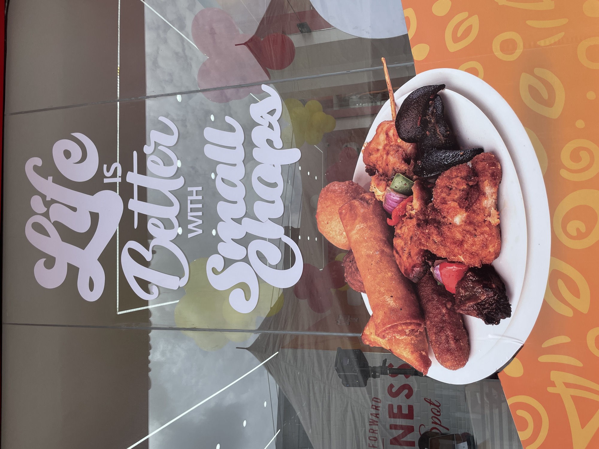 The image shows a close-up of a white plate filled with an assortment of what appears to be fried foods, including spring rolls, chicken pieces, and grilled meat on skewers. The plate sits on an orange surface with a pattern of yellow swirls. Reflected in a glass surface behind the plate is a large vinyl sign that reads "Life is Better with Small Chops" in white lettering. The reflection also shows part of a building exterior and what appears to be a cloudy sky. Based on the sign and food presented, the image likely depicts a food advertisement for "small chops", a popular Nigerian snack platter. Based on the reflection of "Forward Fitness Spot", the location may be in Lekki, Nigeria, known for its fitness facilities and businesses.