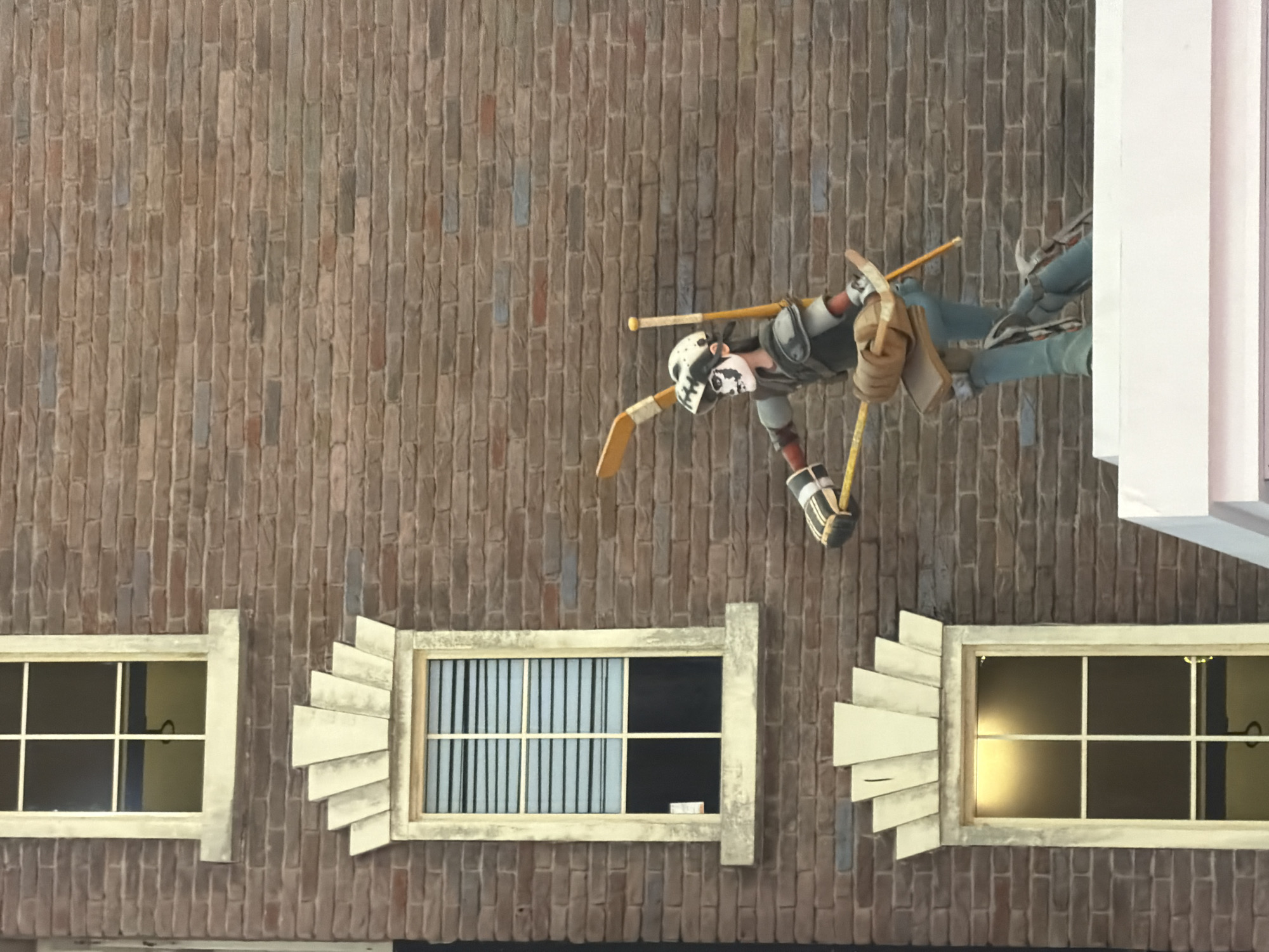 The image depicts a brick building with three windows and a figure dressed as a hockey player. The building's facade is constructed of red and brown bricks. The top window is large and square, with a white frame and panes. The middle window is rectangular and features horizontal blinds. The bottom window is also rectangular and illuminated from within. A decorative stone element with a fan-like design is positioned above the middle window. In front of the middle and bottom windows, a figure resembling a stylized hockey player stands on a white platform. The figure is wearing a helmet with a skull design, a grey and white jersey, and blue pants. The player is holding two hockey sticks, one of which is yellow and appears to be broken. The figure's stance suggests movement or action. The overall scene appears to be an outdoor display or exhibit, possibly in an urban setting.