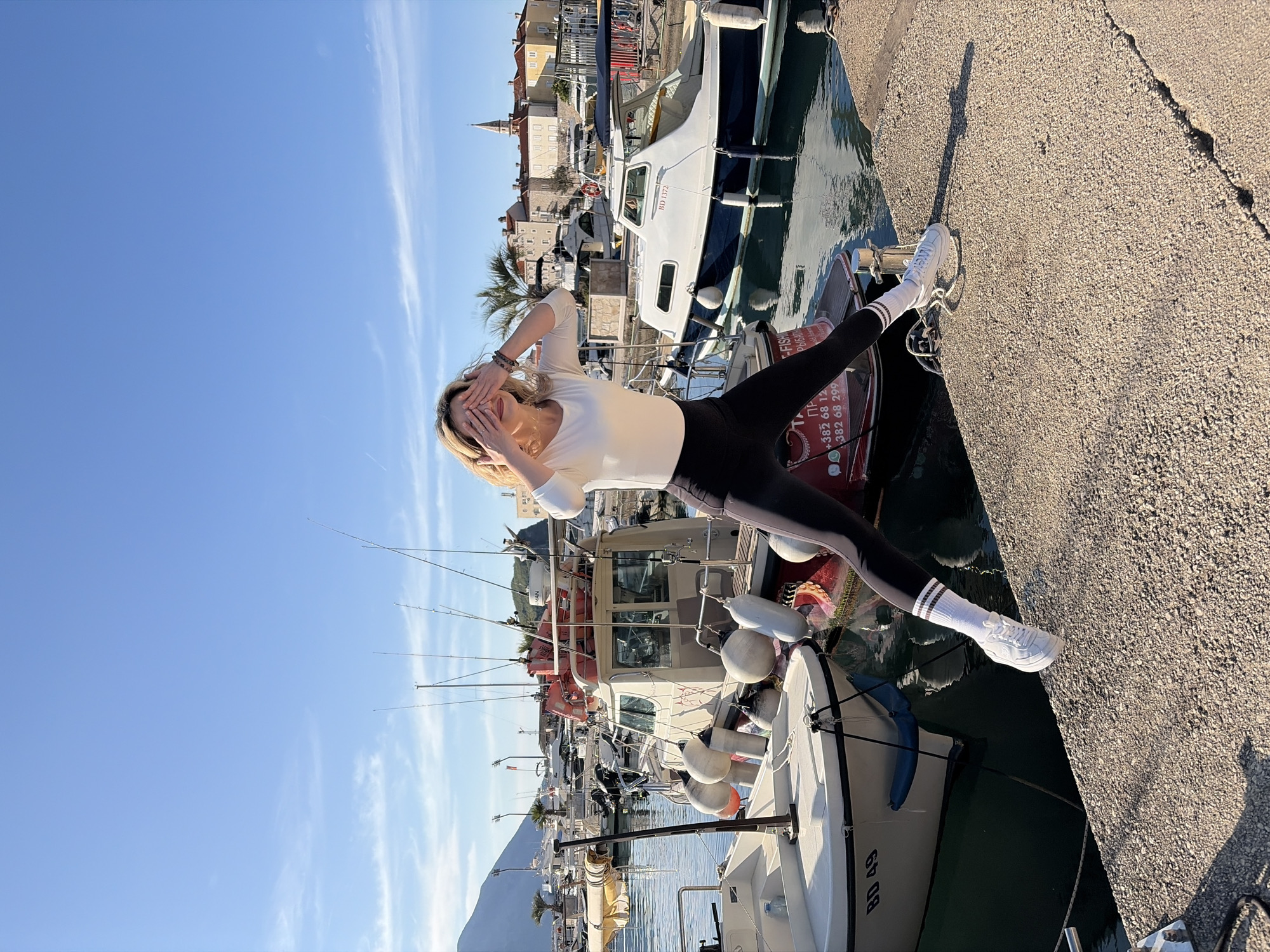 A woman poses for a photograph in Budva, Montenegro. She is positioned with one foot on the concrete edge of a harbor and the other on the side of a boat. She wears a long-sleeved white shirt, black leggings, white socks with stripes, and white sneakers. Her hands are raised to cover her eyes.
The background shows a harbor filled with various boats, with buildings and a church steeple visible in the distance. The sky is blue, and the light suggests it is daytime.