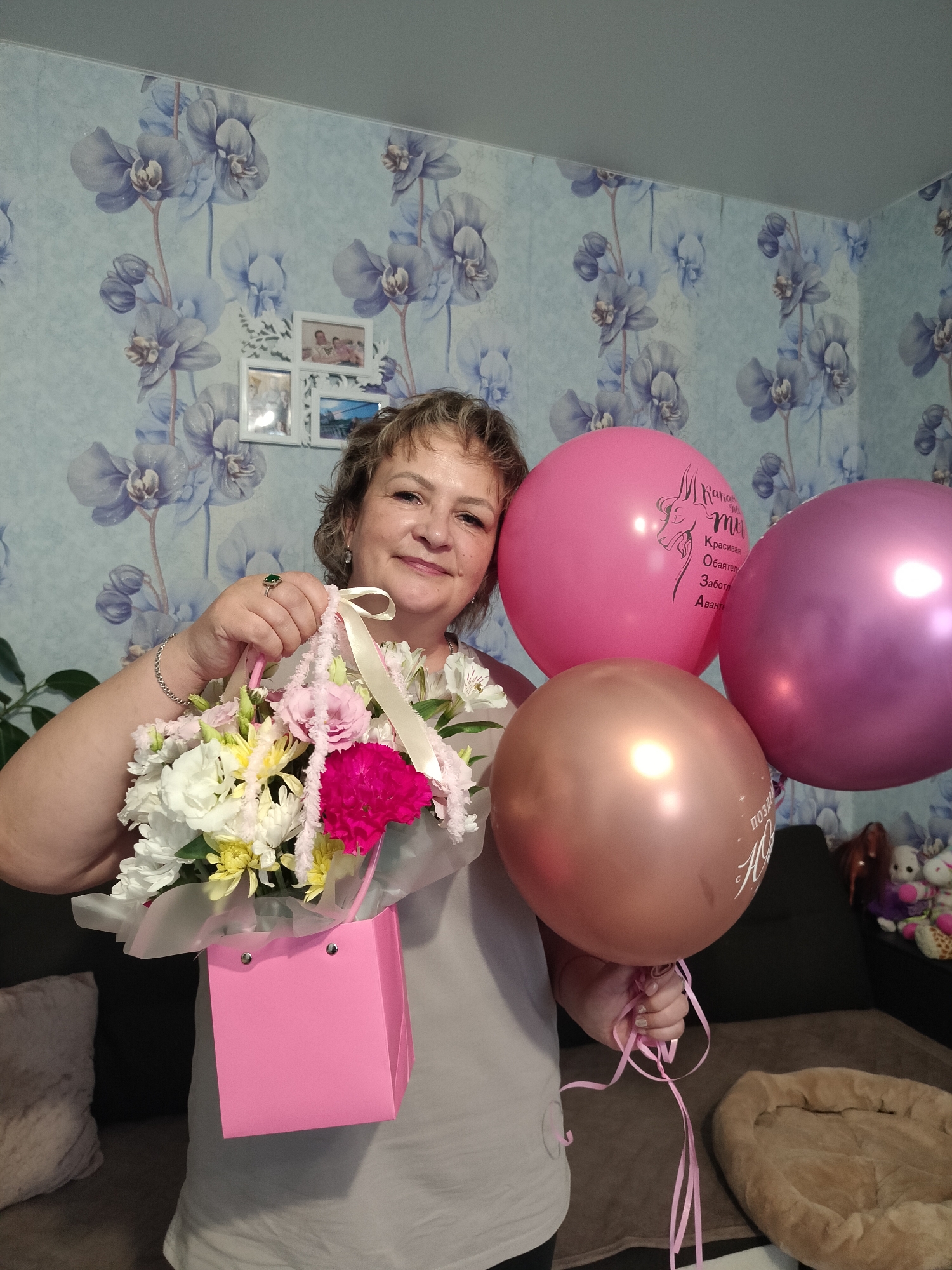 The image captures a middle-aged woman, smiling and holding a bouquet of flowers in a pink gift bag. She is adorned with jewelry, including a ring with a green stone and a bracelet. Three balloons, one pink and two purplish-pink, are visible, with text on two of them. The background features wallpaper with a floral pattern and framed photographs. The scene appears to be an indoor setting, possibly a living room, with furniture like a sofa and a dog bed visible in the periphery. The overall impression is that of a celebration or a special occasion. Given the context provided about the city of Yashkino, Russia, this image likely depicts a moment from a residential environment in that location, perhaps a personal celebration.