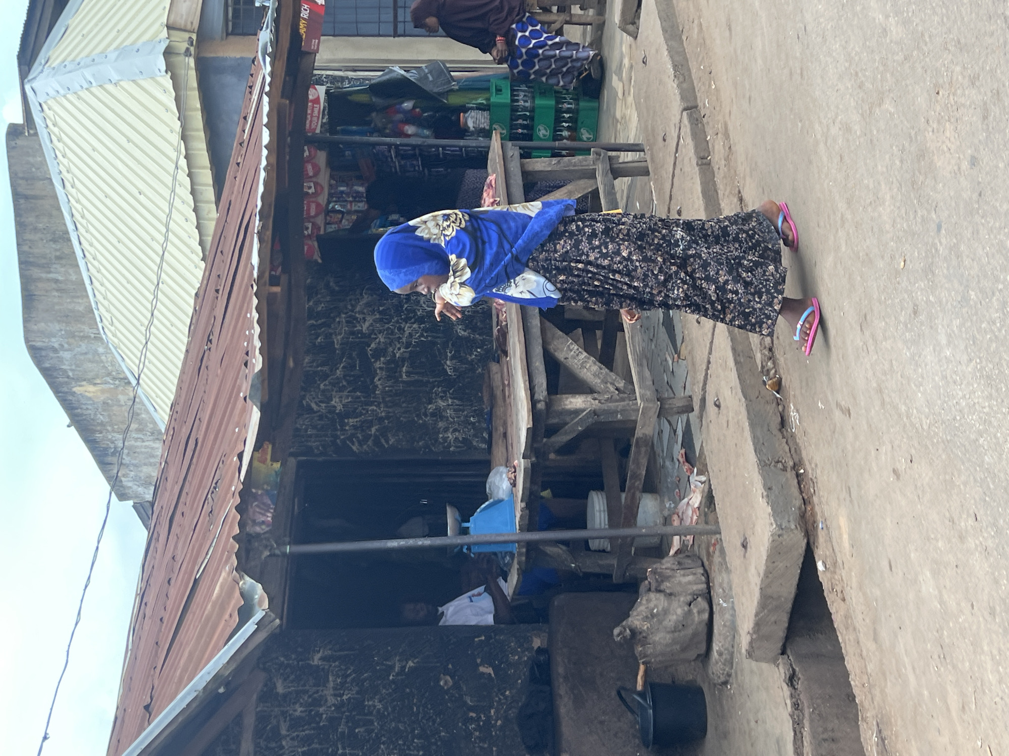 The image captures a street scene in Jos, Nigeria. In the foreground, a young person wearing a blue hijab and a long, floral-patterned skirt walks to the right. They are wearing pink flip-flops. To the left, a wooden market stall with a rusty corrugated iron roof displays various items, including what appears to be meat on a wooden table. A man in a white t-shirt and blue shorts sits behind the table, with a blue scale in front of him. Behind the stall, a woman wearing a dark brown hijab and a patterned skirt sits on a stool. The street is paved and appears to be slightly dirty, with a concrete drain in the foreground. The background shows buildings with metal roofs and weathered walls, and some green crates stacked up. The sky is overcast.