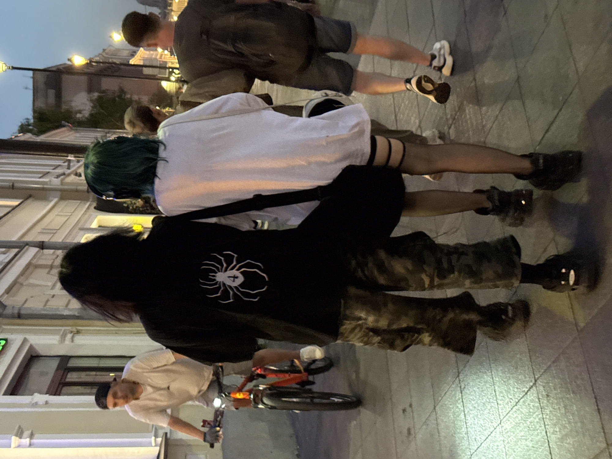 The image depicts a street scene in Moscow, Russia, at dusk. In the foreground, two individuals walk away from the camera. The person on the left has long, dark hair and wears a black t-shirt with a white spider emblem on the back. They are also wearing camouflage pants and dark, chunky shoes. The person on the right has short, green hair and wears a white oversized t-shirt, fishnet stockings with garter straps, and dark shoes. A black bag is slung over the left shoulder of the person with green hair.
Further in the background, a man wearing a white t-shirt and a baseball cap rides a red bicycle. He is on the left side of the frame. To the right of the two central figures, another person in a dark t-shirt and shorts walks away from the camera, carrying a backpack. The street is paved with light-colored tiles, and lampposts with warm yellow lights are visible along the sidewalk. The buildings lining the street are in the background, illuminated by the fading light.