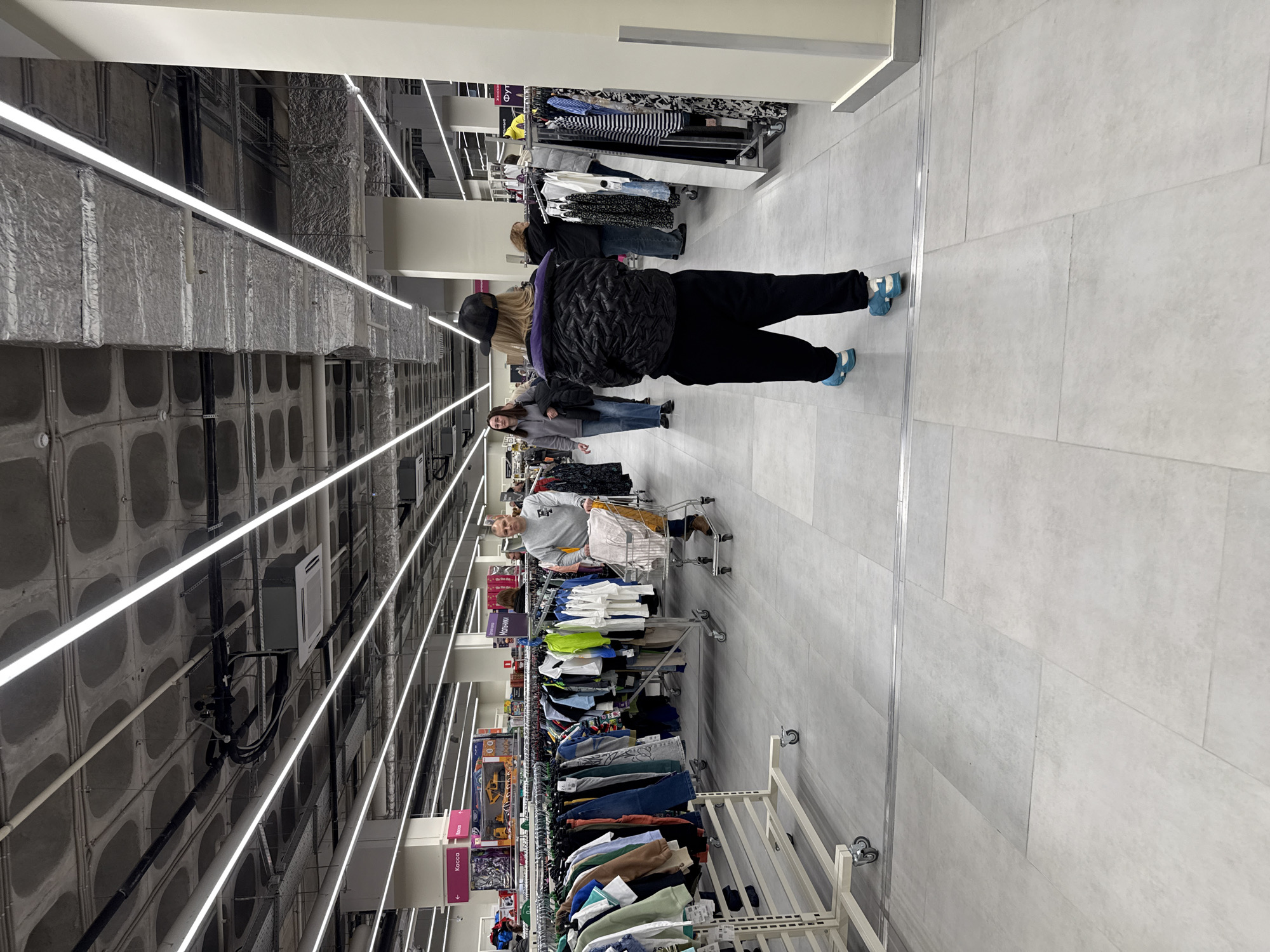 The image shows an interior view of a retail space, potentially a clothing store. The location is identified as Izhevsk, Russia.
The space is illuminated by linear overhead lighting fixtures, creating a modern, industrial aesthetic. The ceiling appears to be unfinished concrete, with visible ducts and pipes. Merchandise is displayed on racks, with clothing items of varying colors and styles.
Several people are visible in the scene, including shoppers browsing the merchandise. One individual is pushing a shopping cart. A woman in a black jacket, black pants, a hat, and blue shoes stands in the center.
The floor is tiled with light gray rectangular tiles. The overall impression is of a functional, well-lit retail environment.