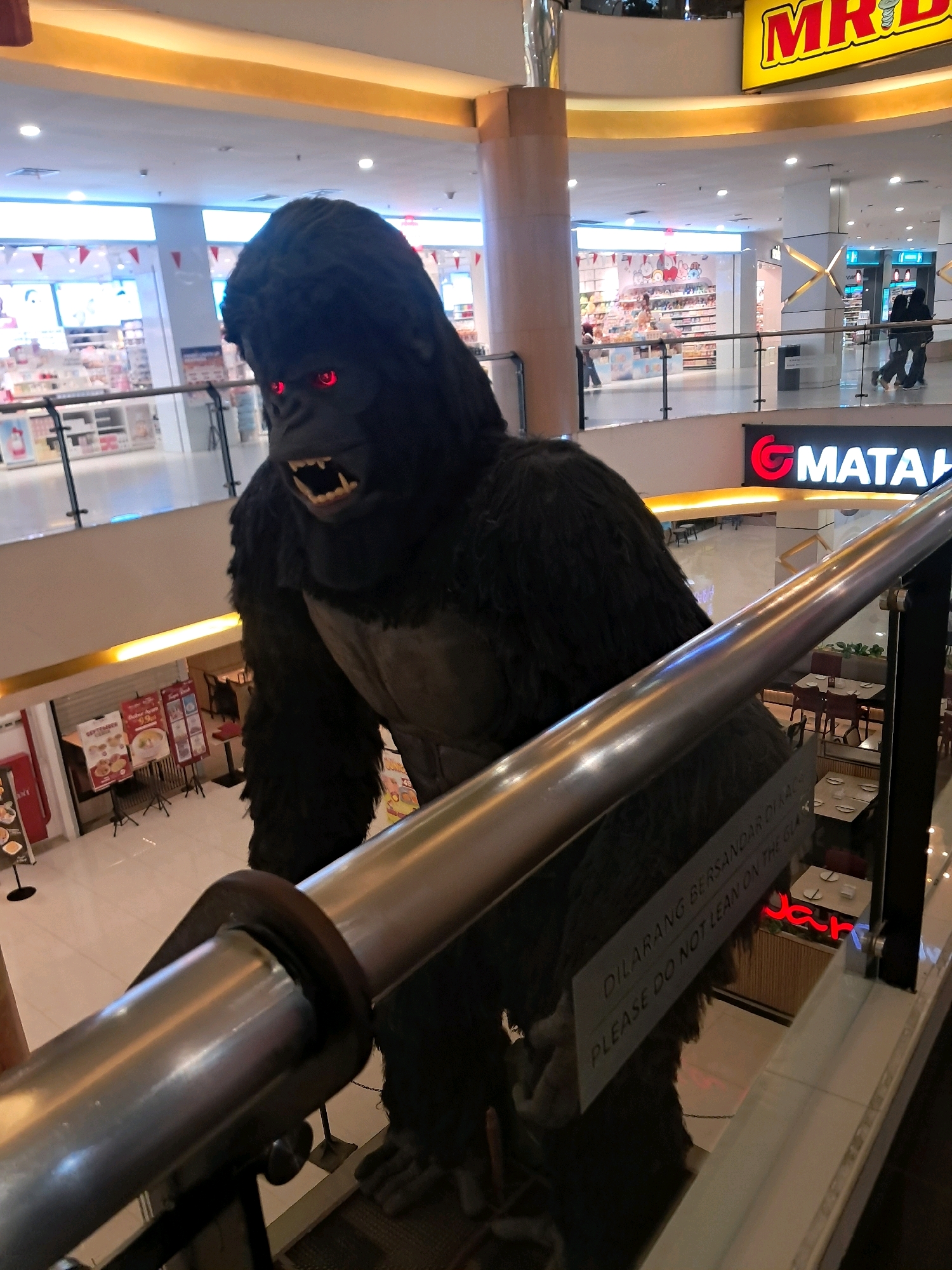 A large, black, furry gorilla statue with glowing red eyes and bared teeth is positioned on an escalator in a shopping mall. The statue is in the foreground, with its body partially obscuring the view of the mall below. The mall appears to be a modern, multi-level structure with shops and escalators visible in the background. There is signage for "MR. D.I.Y." and "MATAHARI" visible on the upper levels. Below the escalator, several tables and chairs suggest a dining area. A sign on the escalator railing reads "DILARANG BERSANDAR DI KACA PLEASE DO NOT LEAN ON THE GLASS." The overall scene depicts a commercial interior in a city.