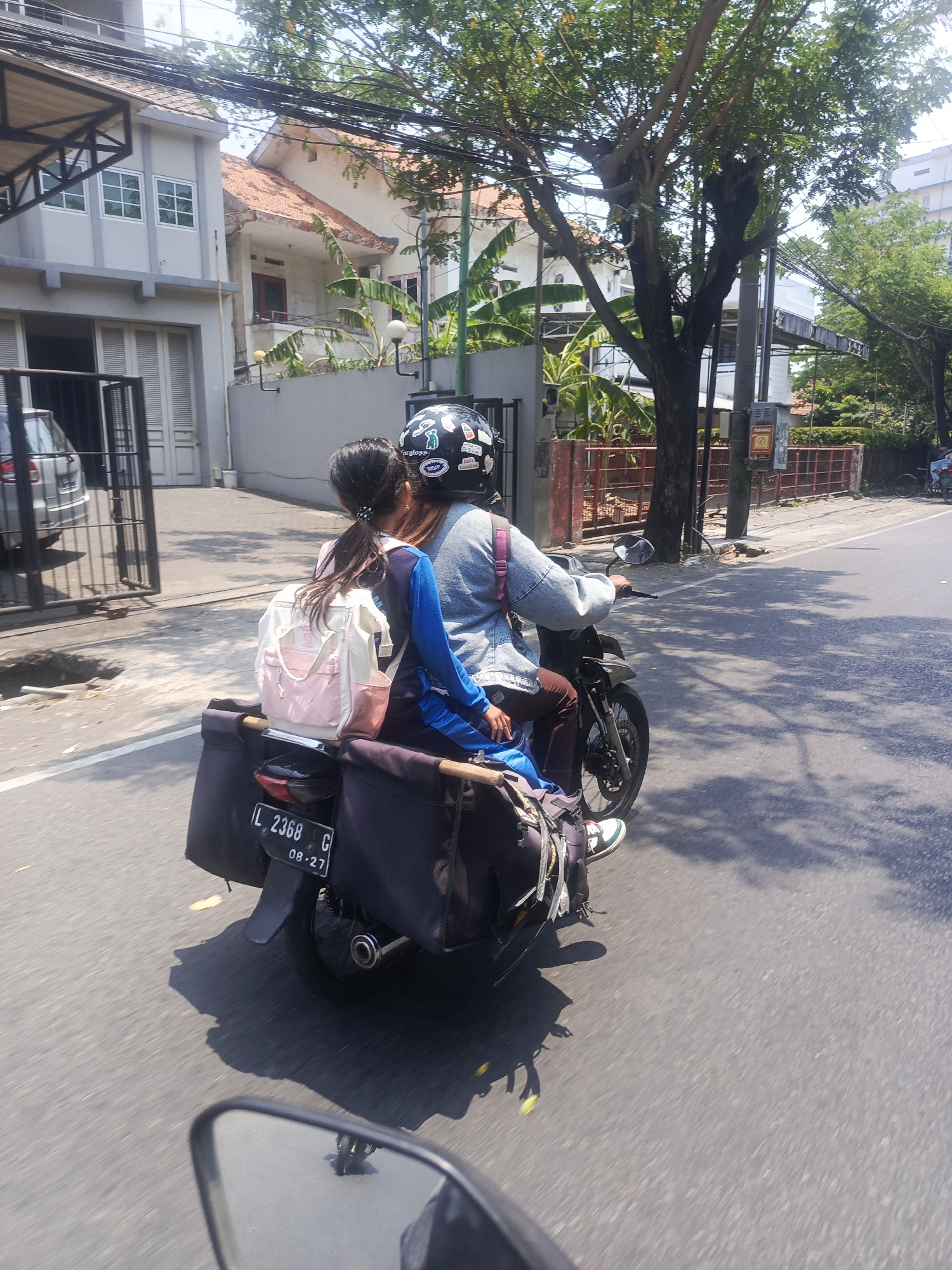 The image shows a rear view of two people riding a motorcycle down a paved road in Surabaya, Indonesia. The driver is wearing a helmet and a light blue jacket, while the passenger, a young girl, is wearing a blue and white shirt and a pink and white backpack. They are both seated on the motorcycle, which is equipped with two large black bags on the rear. The license plate of the motorcycle is visible and reads "L 2368 G 08-27". The background features buildings, trees, and power lines, indicating an urban environment. Sunlight casts shadows on the road, suggesting it is daytime.