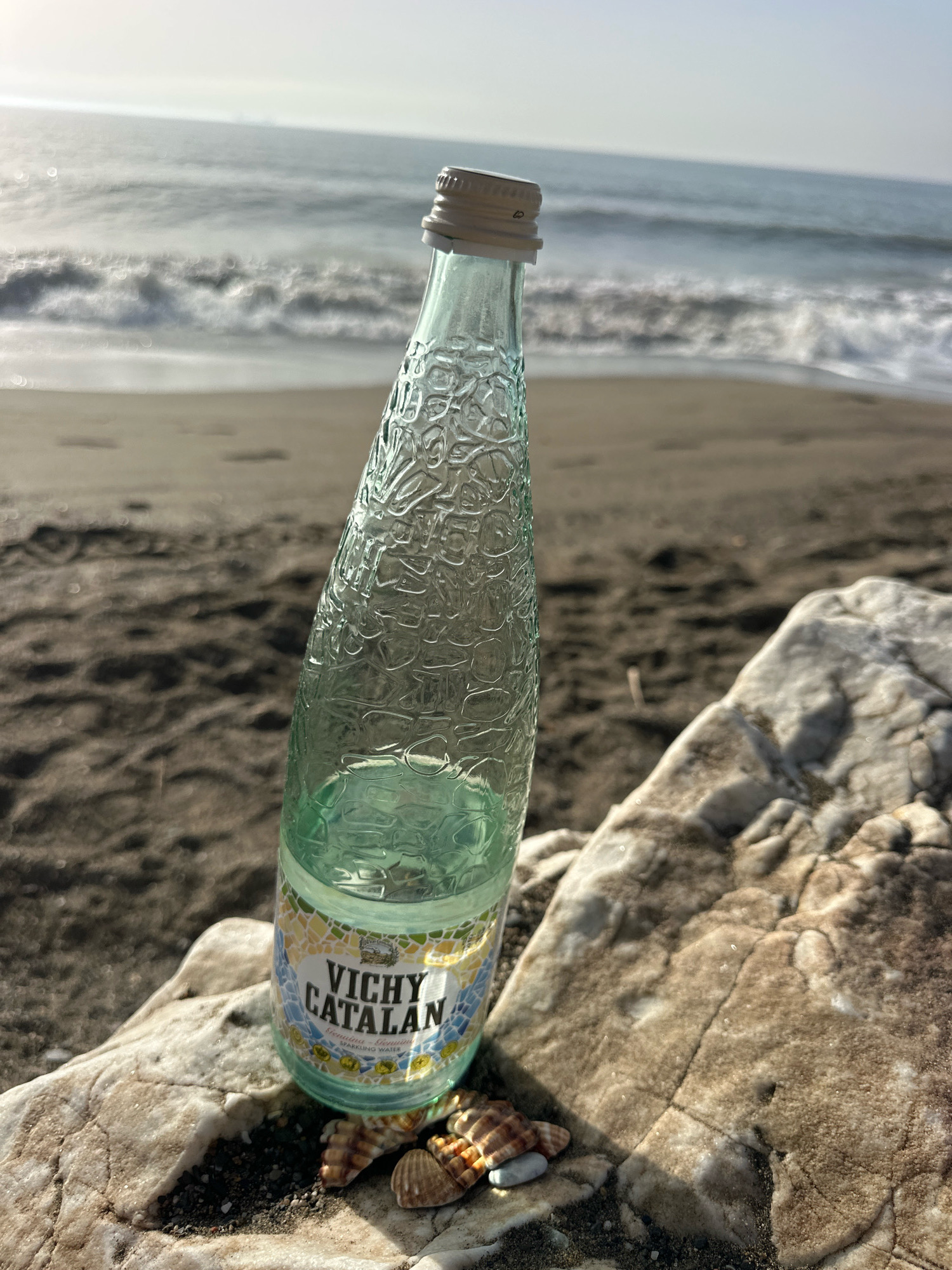A slightly tilted, clear glass bottle of Vichy Catalan sparkling water rests on a rock on a sandy beach. The bottle has a ribbed texture and a silver cap. A label on the bottle reads "VICHY CATALAN" in large, dark letters, with smaller text below. Two small seashells and a gray pebble are positioned near the base of the bottle. The sand on the beach is dark and wet, with small indentations visible. In the background, gentle waves lap against the shore under a hazy sky. The overall impression is one of a serene, coastal setting. The location is not specified in the image or in the prompt.
